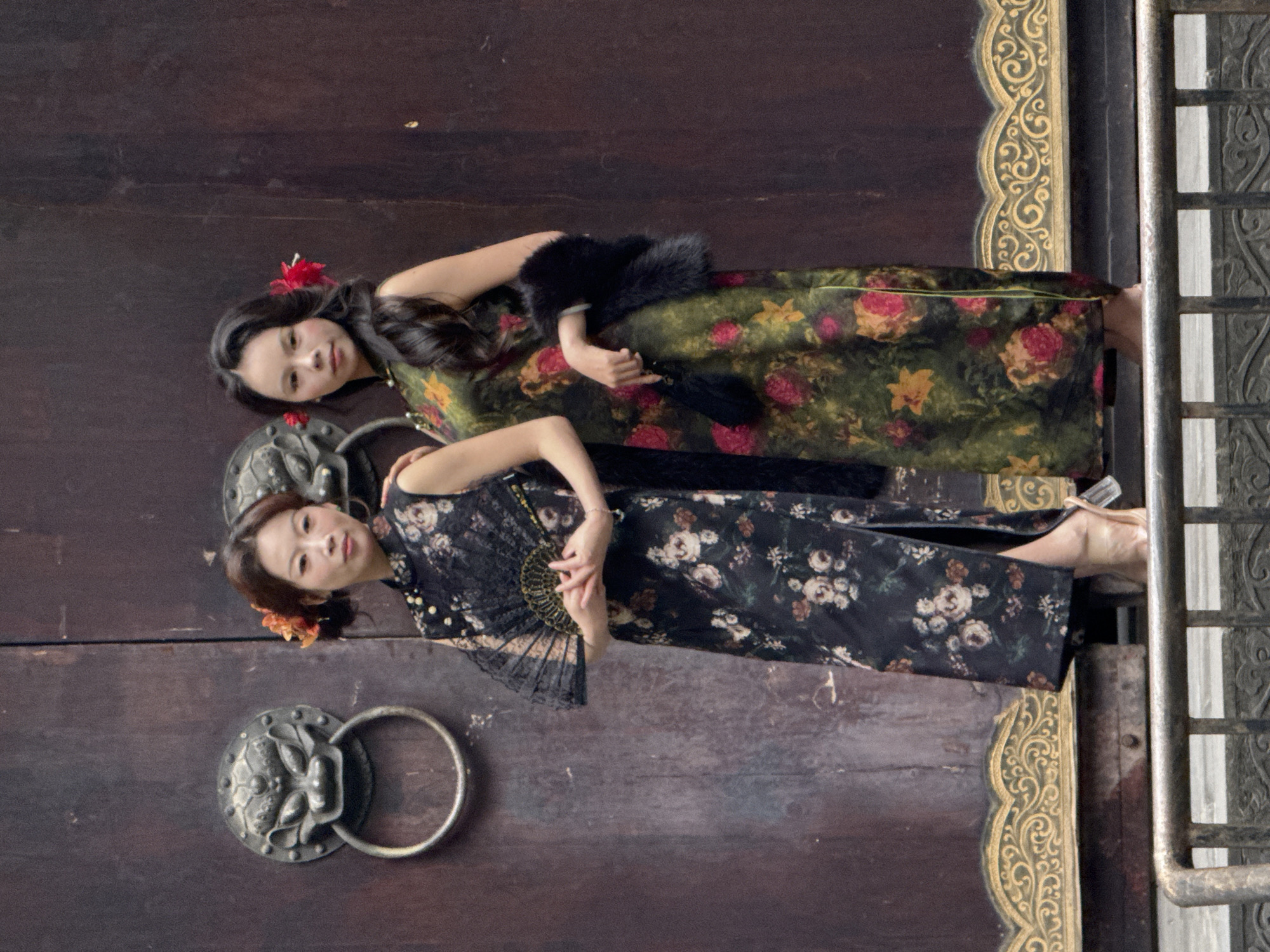 Two Asian women are lying side-by-side on a large, dark wooden door in Yuzhong District, China. The door has decorative metal accents. One woman wears a floral patterned, green and red cheongsam with a black fur stole and a red flower in her hair. She holds a black fan. The second woman wears a black floral cheongsam and has an orange flower in her hair. She also holds a black fan. They are positioned on the lower half of the door, near a lion-head door knocker. The edge of the door features ornate gold trim and is bordered by a dark metal gate.