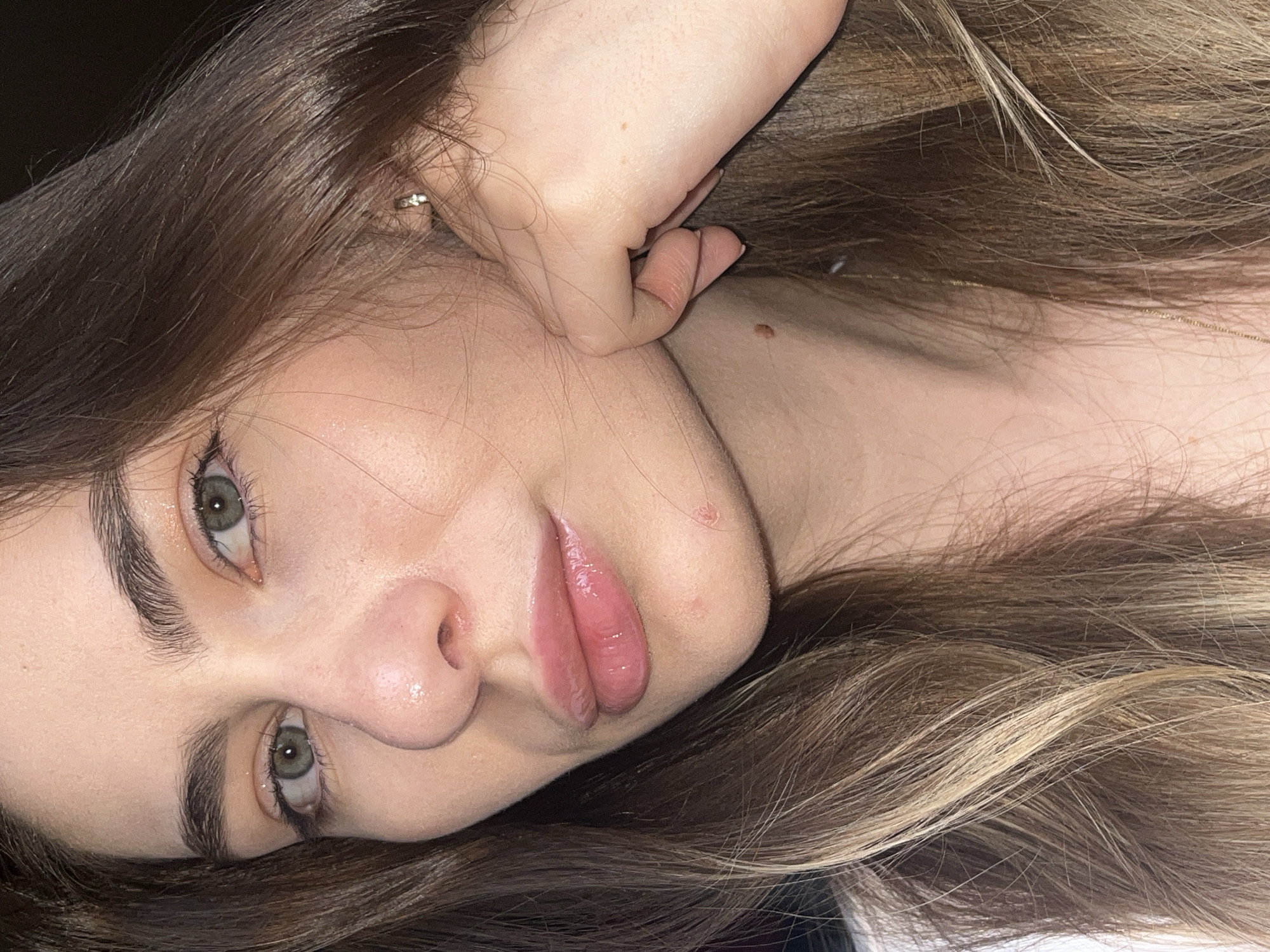 A young woman with green eyes and light brown hair is pictured in a close-up portrait. She is lying on her back with her face turned towards the camera. Her left hand is raised near her face, with her fingers lightly touching her cheek. She has full lips with a glossy appearance. Her skin appears smooth and clear, with a few minor blemishes and a mole visible on her neck. The background is dark and out of focus. A thin gold chain is visible on her neck. Her hair is a mix of dark brown and blonde highlights. No location is discernible from the image.