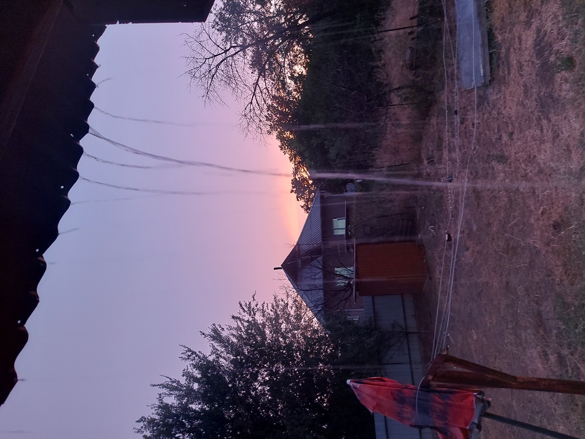 This is an image of a rural scene, likely a dacha or country property in Dachi Su, Kyrgyzstan. The perspective is from under a dark, corrugated overhang, possibly a roof or porch. Several thin streams of water are falling from the edge of this overhang, suggesting it might be raining, or perhaps sprinklers are in use.
The background depicts a twilight sky with hues of soft purple and pink, indicating either dawn or dusk. Silhouetted against the sky are the bare branches of a tree on the left and the dense foliage of trees and bushes on the right. In the mid-ground, a rustic building is visible. It has a dark roof and what appears to be a reddish-brown section, possibly an extension or shed. Windows on the building are dimly lit. A fence runs along the property line.
The ground is covered in dry, straw-like grass. In the foreground, on the right side, a partially unfurled red and black patterned umbrella or tarp is propped up. The overall mood of the image is tranquil and slightly melancholic, typical of the quiet moments at the end or beginning of the day in a rural setting. There is no visible text in the image.