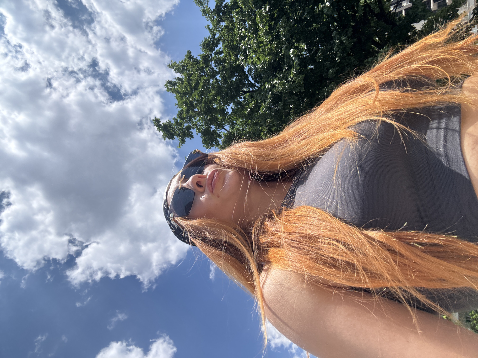 A low-angle, full-body shot captures a woman with long, vibrant orange hair, wearing sunglasses and a black bandana. She is looking upwards, with her mouth slightly puckered. She is dressed in a dark grey tank top. The background is a bright blue sky with fluffy white clouds, and a lush green tree. The scene appears to be outdoors in a city, possibly Budva, Montenegro, given the natural light and common foliage found in the region. The shot is taken from a low perspective, emphasizing the sky and the woman's upward gaze.