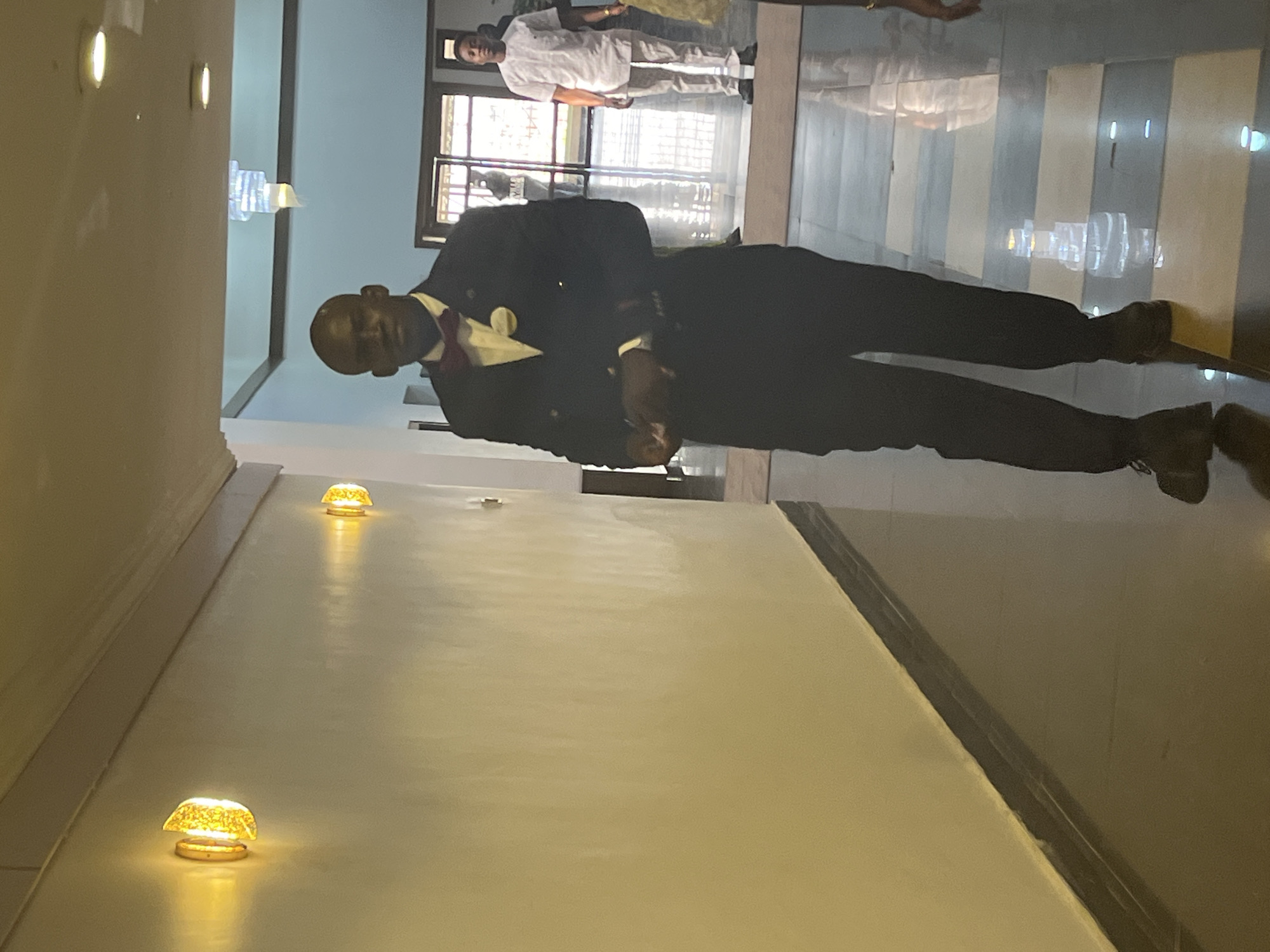 A man in a suit stands inside a building in Abuja, Nigeria. He is dressed in a dark suit with a lighter-colored dress shirt and a bowtie. Another man dressed in lighter-colored clothing can be seen in the background, near what appears to be an exterior window or doorway. The building's interior features tiled floors and lights mounted on the walls.