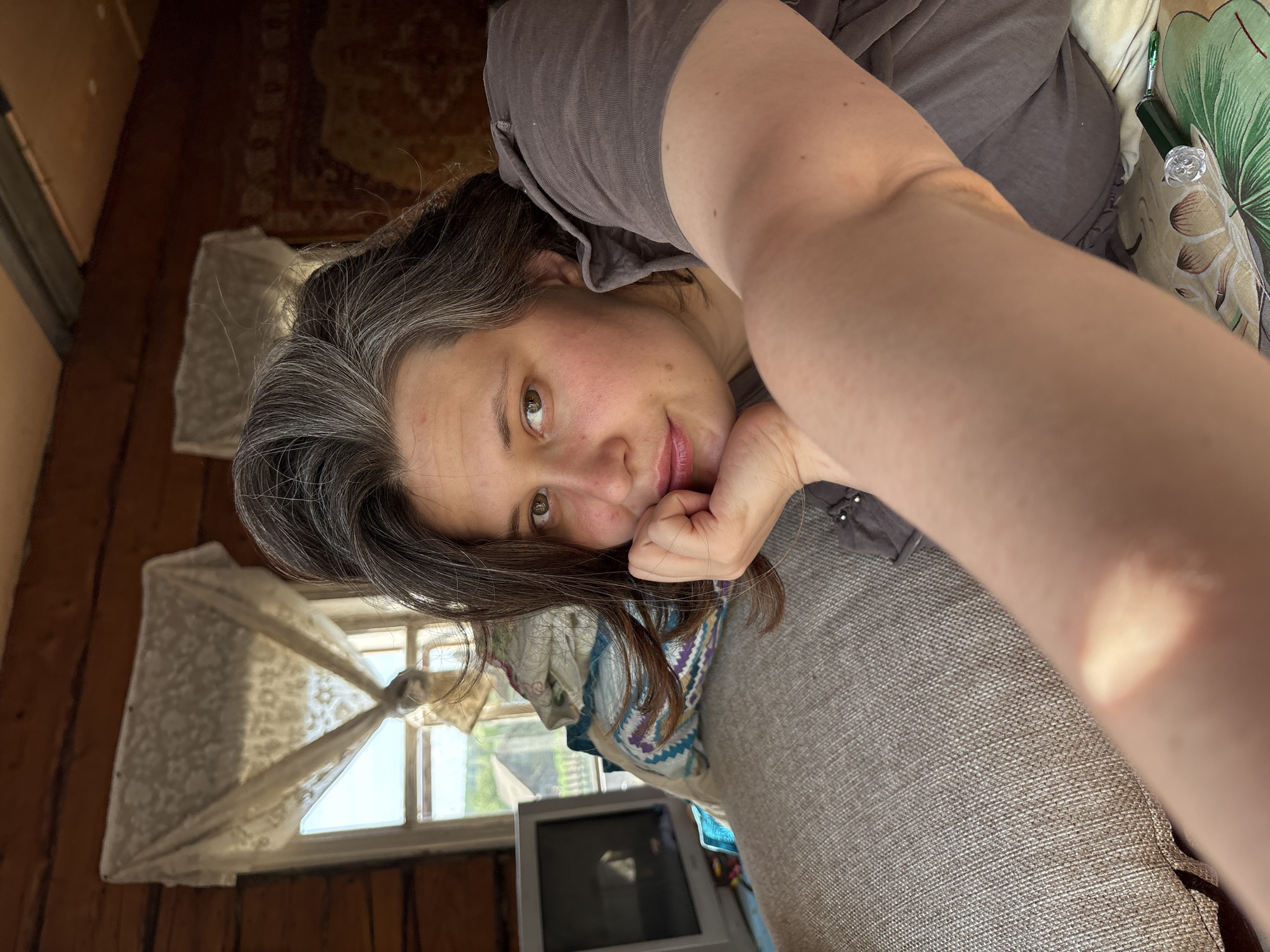 A woman with grey hair is lying on a grey couch and resting her head on her hand. She is wearing a brown t-shirt. A window with a white, lace curtain is behind her. A television is visible in front of the window. The location is Nizhnyaya Chermoda, Russia.