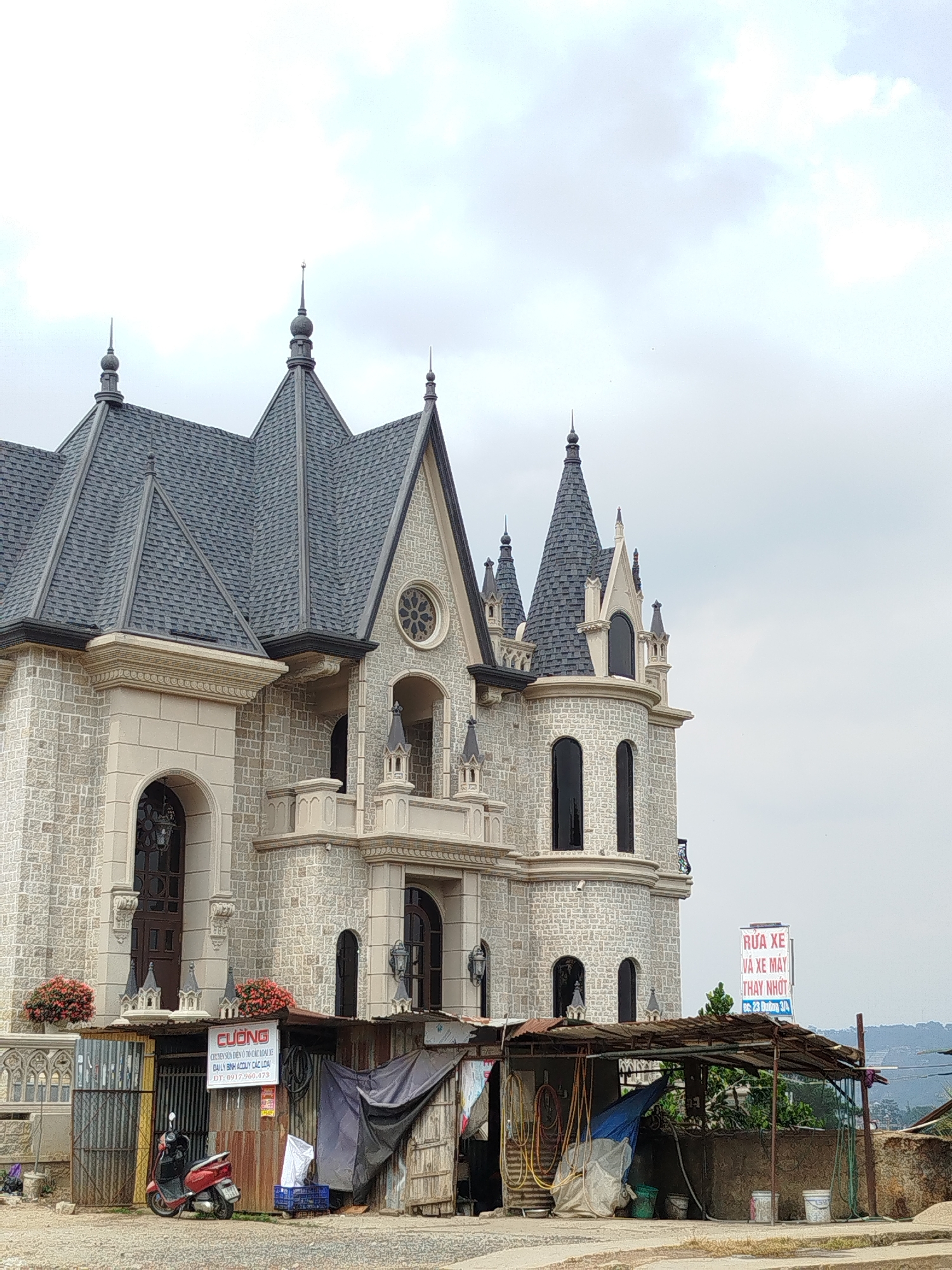 An eye-level shot captures a striking juxtaposition in Da Lat, Vietnam, featuring an ornate, castle-like building towering over a humble, makeshift structure in the foreground. The grand building, constructed with light-colored stone blocks, boasts multiple pointed grey slate roofs, turrets, and arched windows, giving it a distinct European architectural feel. Intricate details like small decorative spires, ornate balconies, and a circular stained-glass window (or similar design) adorn its facade.
In stark contrast, immediately in front of this elaborate building is a utilitarian shanty, constructed from corrugated metal sheets and draped tarps, serving as a roadside business. A red scooter is parked to the left of the shanty, indicating local activity. Various hoses, buckets, and other repair-related items are visible around and under the shelter of the shanty.
The scene appears to be set on an overcast day, with a muted grey and white sky dominating the upper portion of the image. Distant green hills are faintly visible on the right horizon, suggesting a somewhat elevated or rural setting.
Visible text on a sign attached to the shanty reads: "CƯỜNG", "CHUYÊN SỬA ĐIỆN Ô TÔ CÁC LOẠI XE", "ĐẠI LÝ BÌNH ACQUY CÁC LOẠI", "ĐT: 0917.960.473," indicating a car electrical repair and battery dealership. Further to the right, a larger sign advertises: "RỬA XE VÀ XE MÁY THAY NHỚT ĐC: 23 Đường 3/4," identifying a car and motorbike wash and oil change service at address 23 Duong 3/4. No people are visible in the frame.