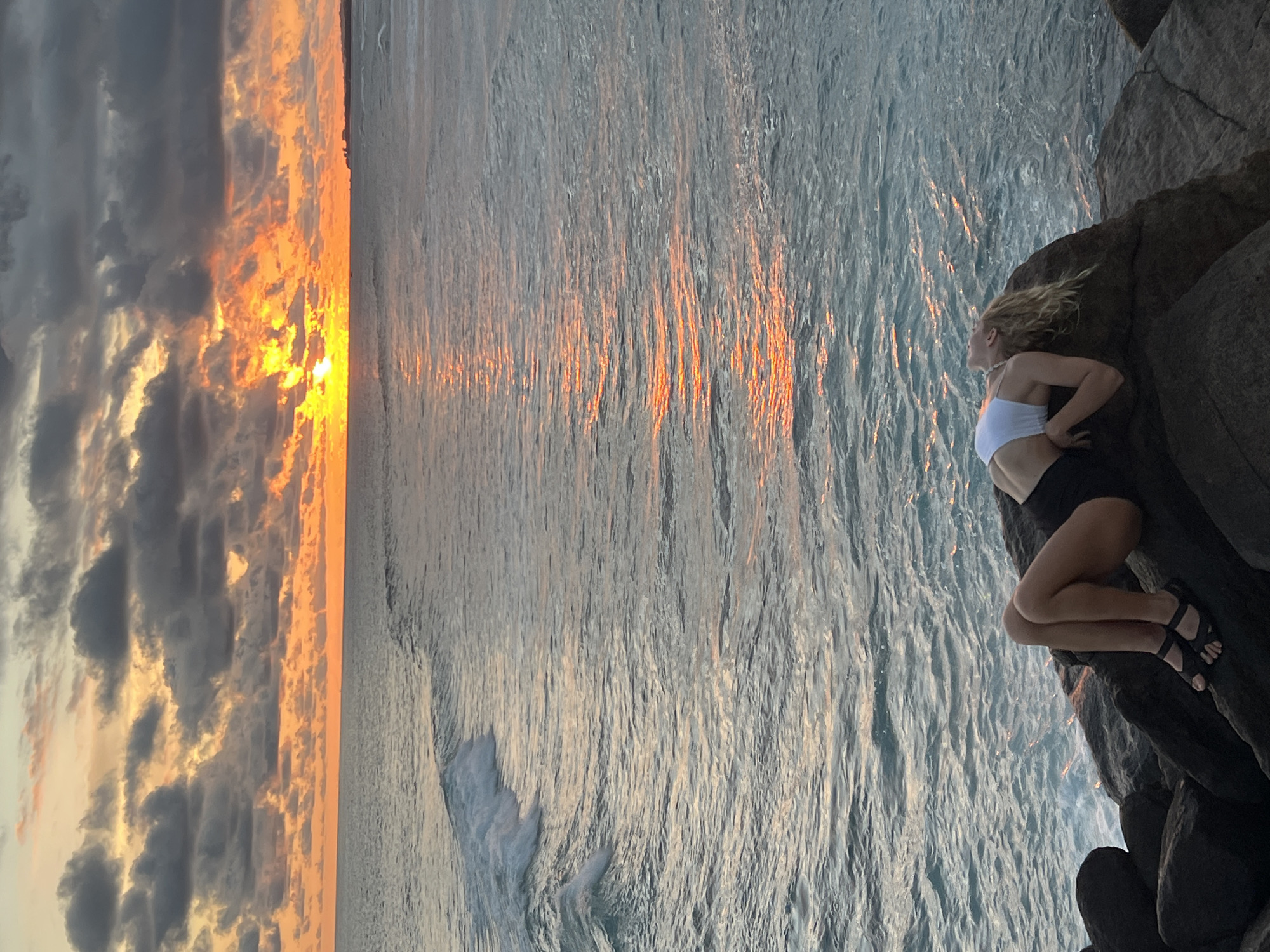 A woman is sitting on dark rocks overlooking the ocean at sunset. She is wearing a white top, dark shorts, and black sandals. The sunset is visible in the sky, with the sun's reflection shimmering on the water. The ocean appears wavy. The city of Goviyapana, Sri Lanka, is not discernible from this image.