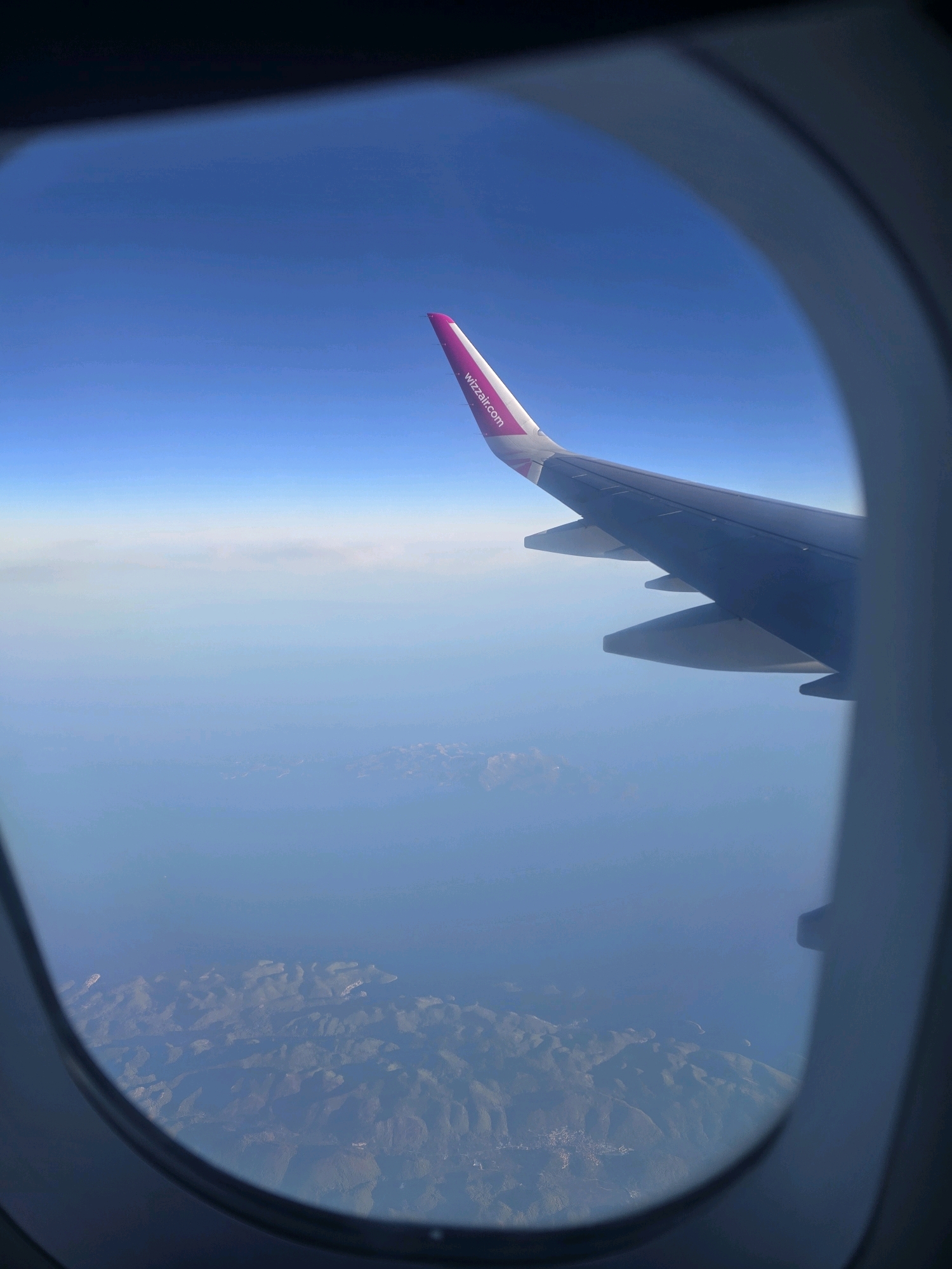 The image is taken from an airplane window, looking out at the sky and land below. The wing of the airplane is visible in the foreground, with the logo "wizzair.com" printed on it in pink. Below the wing, a vast expanse of blue sea stretches out, dotted with islands or coastal landmasses. The land appears mountainous and rugged, with some patches of what could be vegetation or settlements visible. The sky is a clear, vibrant blue. The curved edge of the airplane window frames the entire scene. While the image captures a scenic aerial view of a coastline, it does not provide specific enough detail to definitively identify the location as Zavala, Croatia. The distant land formations are not distinct enough to pinpoint a specific city or town.