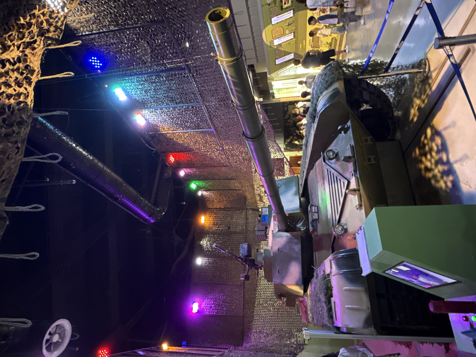 The image shows a tank on display at what appears to be a museum. The tank is surrounded by netting and colorful stage lighting, primarily blue, red, green, yellow, white and purple. The background features a crowd of people. The location of the scene is 江北城街道, China.