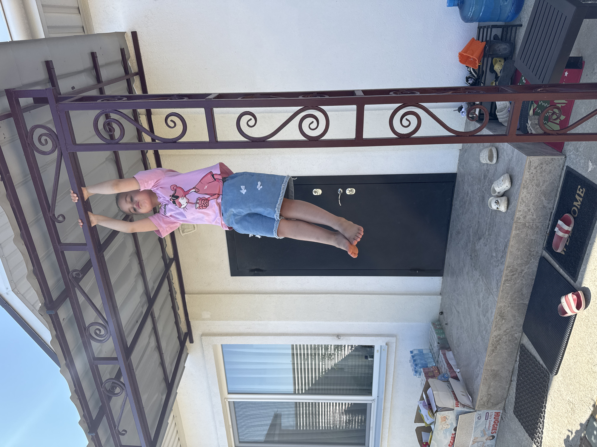 A young child is hanging from a metal bar attached to the porch of a building, with their feet in the air. The child is wearing a pink t-shirt with a cartoon character and blue denim shorts. The building appears to be a single-story structure with a white exterior wall and a dark door. In front of the building, there is a concrete porch with some boxes and water bottles stacked on it. There are also several pairs of shoes scattered on the porch and the ground in front. The scene is outdoors, under a clear blue sky, suggesting a sunny day. This image was captured in Yevpatoriya, Ukraine.