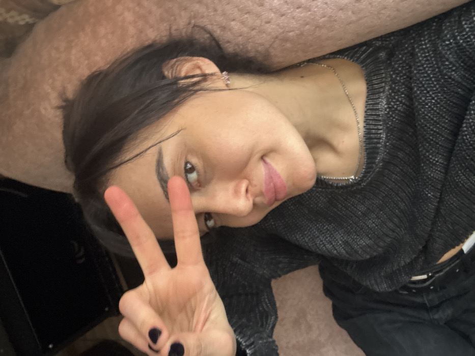 A female subject is depicted in a semi-reclined position, directly facing the camera with a slight smile. Her left hand is raised, forming a V-sign with the index and middle fingers, positioned partially over her left eye. She wears a dark grey ribbed sweater, revealing a thin silver-colored chain necklace. Small, light-colored earrings are visible, and her fingernails are painted dark. Her hair is dark brown. The background features a dark, angular object on the upper left and a light brown, textured surface on the right. The scene is located in the city of Bolnisi, Georgia.