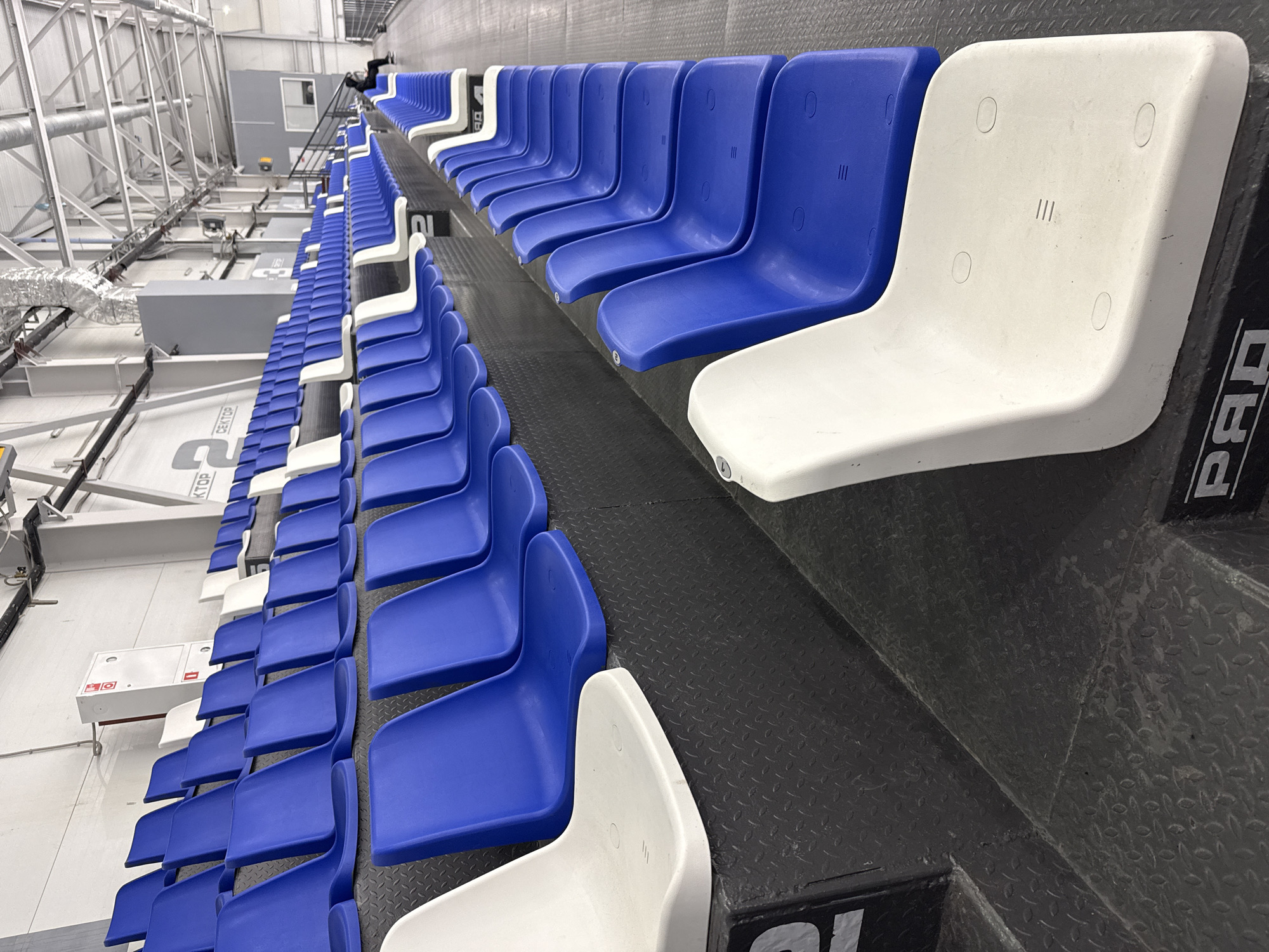 The image depicts rows of stadium seating in an indoor venue, likely in Votkinsk, Russia. The seats are alternating blue and white. The seating platform has a dark gray diamond plate surface. Overhead are structural elements and ductwork. No people are visible.