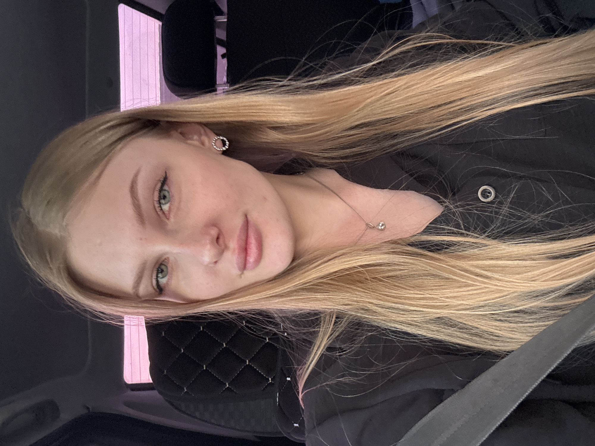 A fair-skinned woman with long blonde hair, wearing a black top and a necklace, is in a vehicle. The interior of the vehicle is dark, with a quilted headrest visible and a seatbelt across the person. A building with a pink facade is visible through a window in the background. The location is Кондратово, Russia.