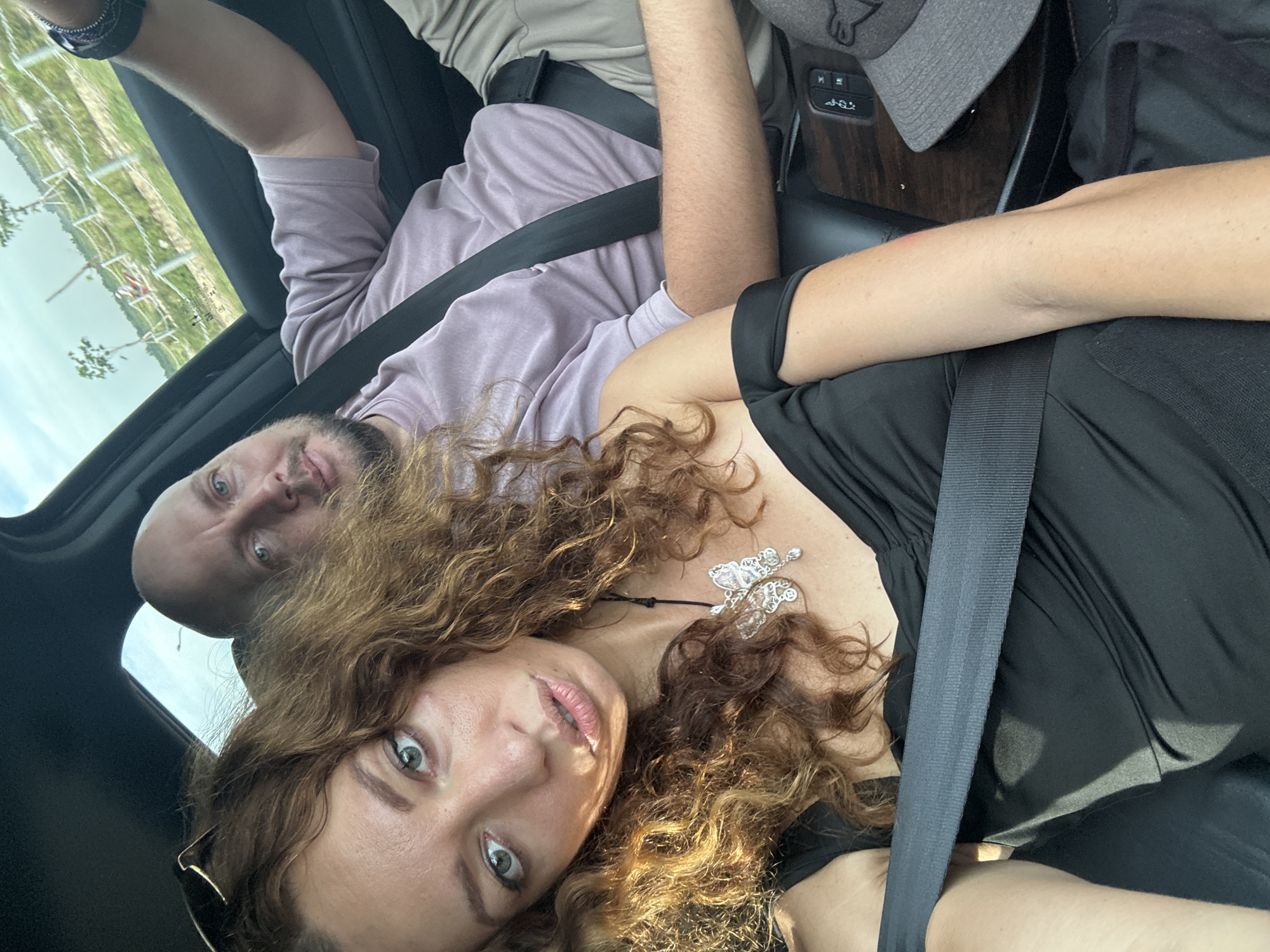 The image shows two individuals, a woman and a man, inside a car. The woman is in the foreground, taking the selfie, with her curly brown hair framing her face. She is wearing a black off-the-shoulder dress and a butterfly-shaped pendant necklace. The man is in the background, in the passenger seat, wearing a light purple t-shirt. Both individuals are wearing seatbelts. The car appears to be in motion, with a view of trees and a white fence visible through the car window, suggesting an outdoor environment, possibly in a suburban or rural area. The scene does not provide specific visual cues to definitively pinpoint the location within Hoa Lư Ward, Vietnam.