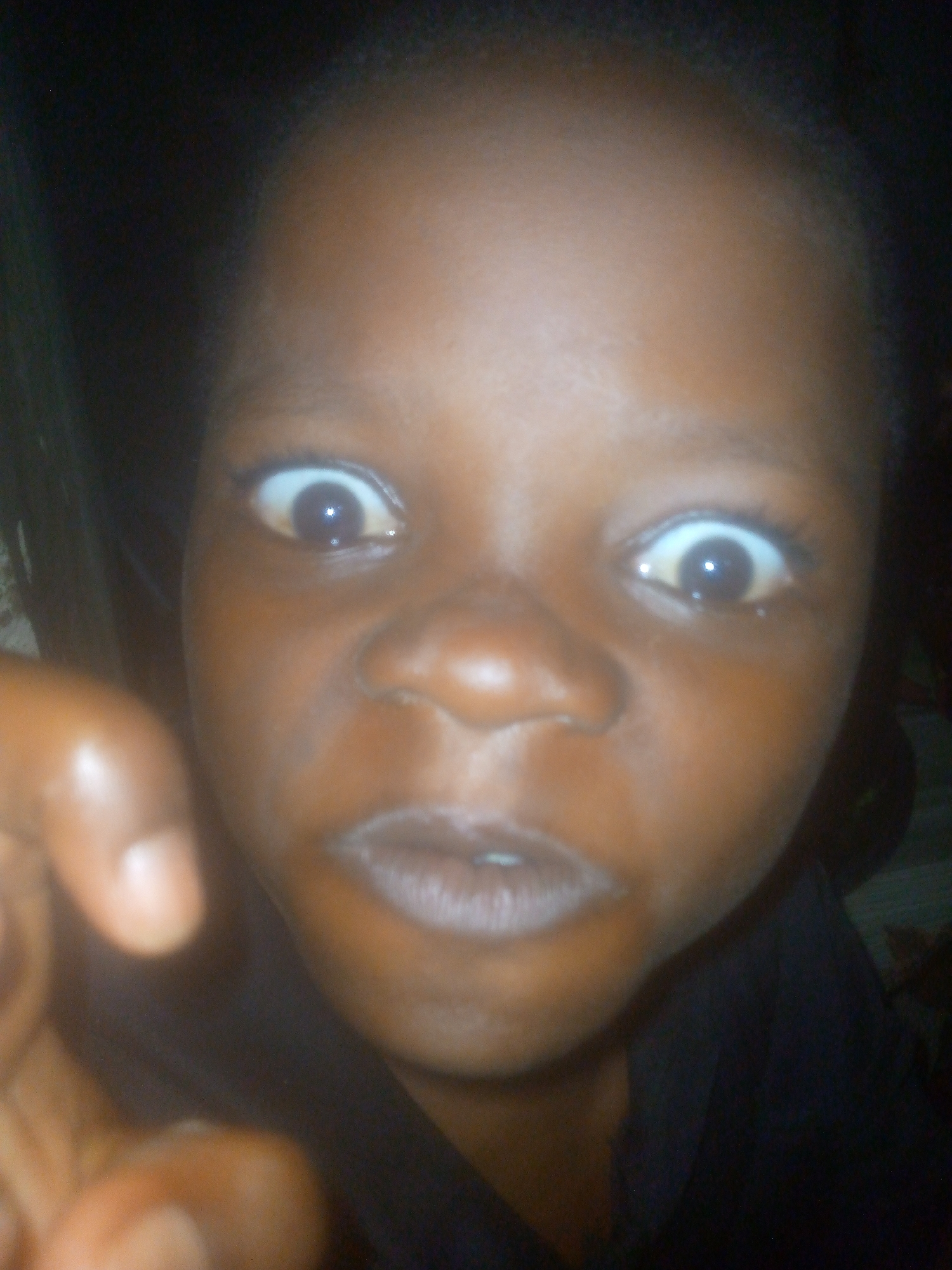 A close-up, slightly blurry image depicts a child with brown skin, wide eyes, and an open mouth, suggestive of surprise or shock. Part of the child's hand is visible in the lower left corner. The background is dark and indistinct, preventing any identification of the location within Ikorodu, Nigeria.