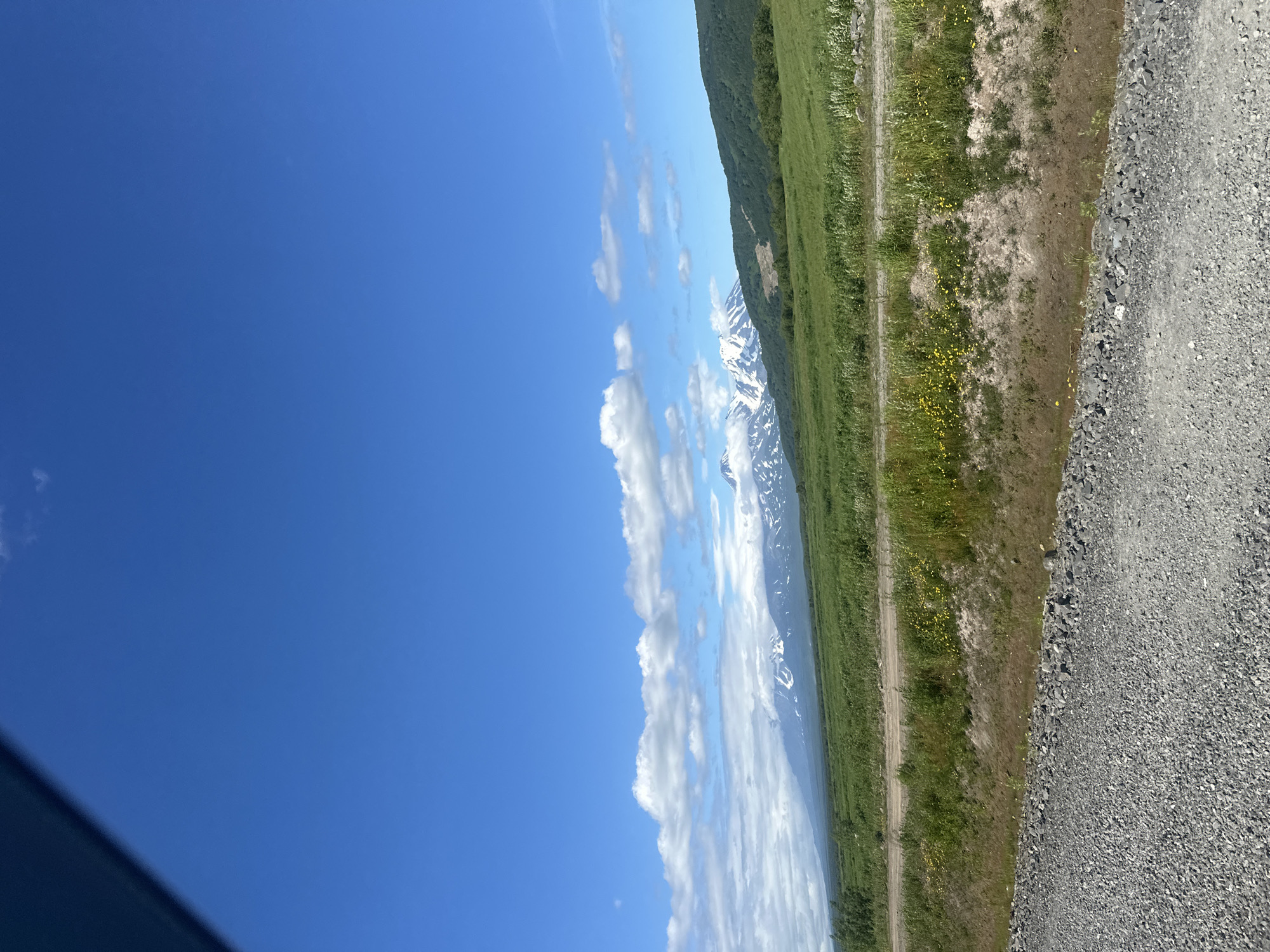 The image depicts a wide, natural landscape under a bright blue sky with scattered white clouds. In the distance, a mountain range with snow-capped peaks is visible, some partially obscured by clouds. The foreground features a gravel road or path, with sparse vegetation and some yellow wildflowers growing along its edge. Beyond the immediate foreground, a vast, green, rolling field extends towards the mountains. The scene appears to be a rural or natural area, possibly on the outskirts of Petropavlovsk-Kamchatsky, Russia, given the presence of volcanic mountains common to the region. There are no people or animals visible in the image. The overall impression is one of a serene, expansive natural environment.