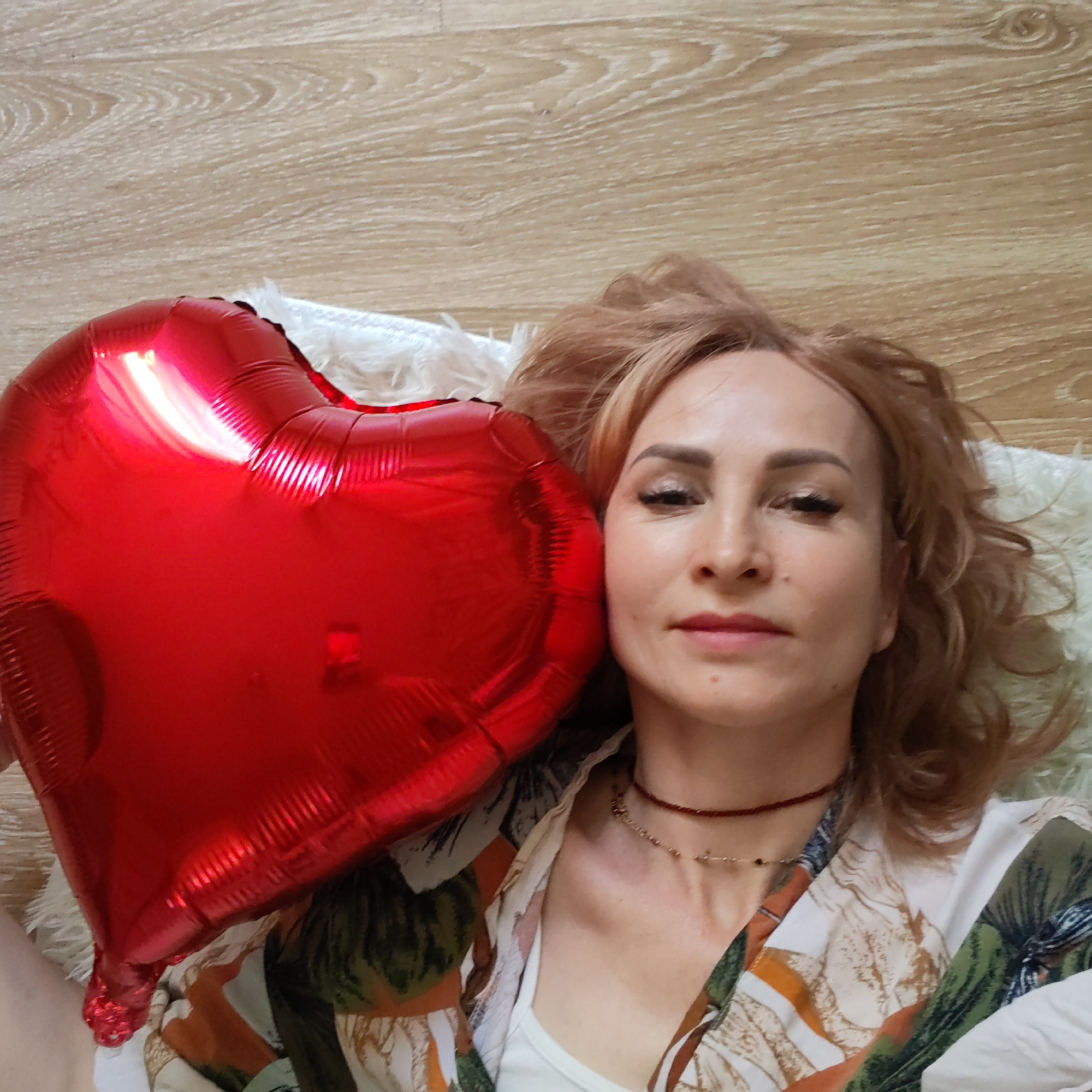 A fair-skinned woman with reddish-blonde hair lies on what appears to be a wooden floor in an indoor setting. She is holding a large, red heart-shaped balloon next to her face. The woman is wearing a printed shirt over a white top, and a pair of delicate necklaces are visible around her neck. The background also includes a small white rug or cushion. It is not possible to confirm the location as the city of Chaikovsky, Russia, solely based on the visual information in the image.