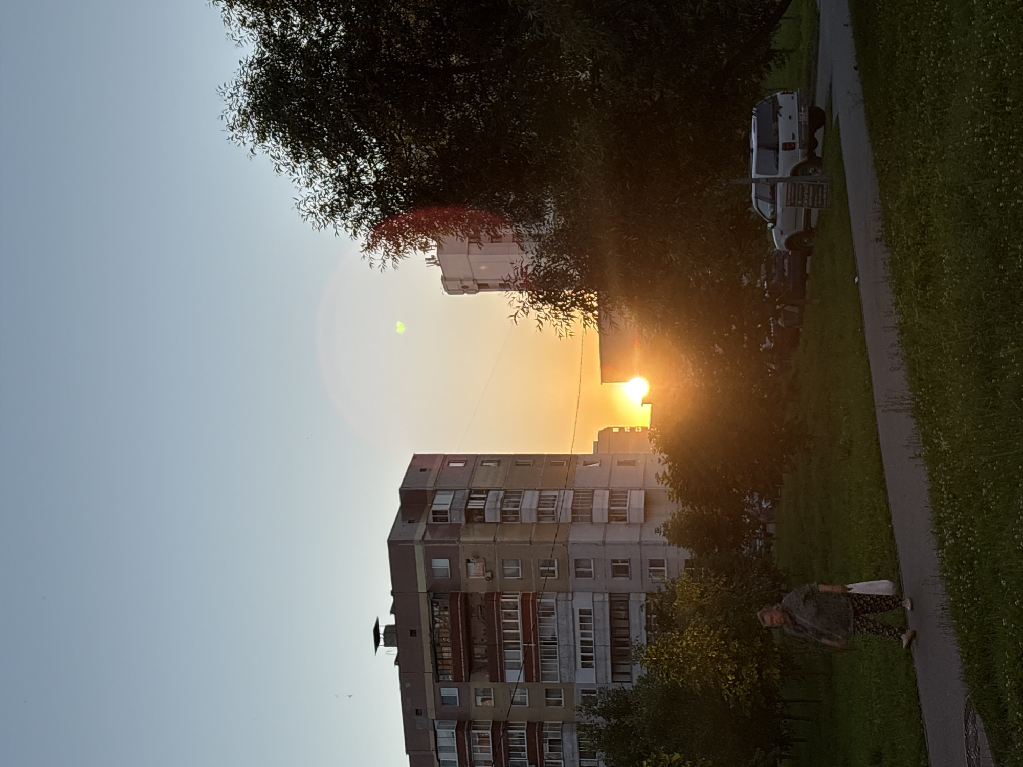 The image captures a sunset in a residential area of Vitsebsk, Belarus. In the foreground, a woman walks on a paved path, carrying a white bag. She is dressed in a green patterned shirt and floral pants. To her left, lush green grass dotted with small white flowers covers the ground. Behind her, trees with dense foliage create a backdrop, with some saplings visible in the mid-ground.
Dominating the mid-ground are two apartment buildings. The one on the left is a multi-story structure with a predominantly beige and gray facade, accented with red and white window frames and balconies. The building on the right, further in the distance, is a lighter colored, taller apartment block.
The setting sun is a prominent feature, appearing as a bright, orb partially obscured by trees and buildings, casting a warm, golden light across the scene. The sky is a clear, pale blue, transitioning to warmer hues near the horizon. In the far background, a white SUV is parked on the right side of the path.
