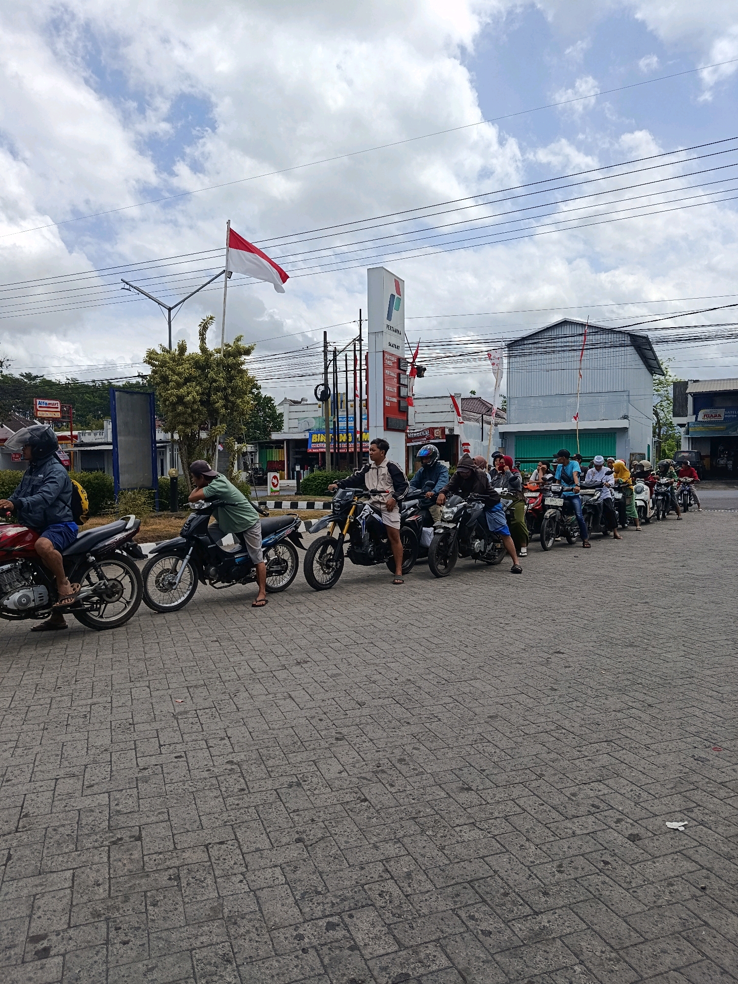 The image shows a line of motorcycles parked on a brick-paved road in Tempeh Lor, Indonesia. The scene appears to be at a gas station, indicated by the prominent "Pertamina" signage and a red and white Indonesian flag flying overhead. Several people are on or near their motorcycles, suggesting they are either waiting in line or have just arrived. The sky is cloudy, and the overall lighting suggests it is daytime. The surrounding area includes buildings that appear to be shops or businesses. The individuals are dressed in casual clothing, with some wearing helmets. The motorcycles are diverse in type and color, and some are parked quite close to each other. The ground is paved with rectangular bricks laid in a herringbone pattern. There are also electrical wires visible overhead, along with a street lamp.