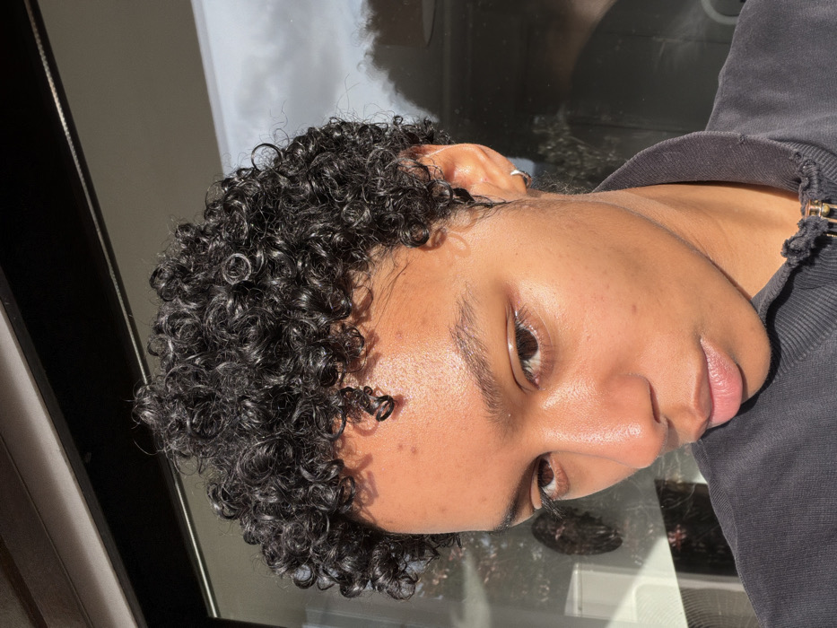 A close-up portrait shot depicts one individual with dark skin and short, dark, tightly curled hair. The person is positioned slightly off-center, facing towards the left, with their gaze directed at the camera. They wear a dark, possibly black, zippered garment, with a gold-colored zipper pull visible at the bottom of the frame. A small silver hoop earring is present in the visible right ear. Strong natural light, likely sunlight, illuminates the left side of the subject's face, highlighting facial features and creating shadows on the right side. The background consists of a blurred reflection on a window or glass surface, showing a bright, partially cloudy sky and indistinct dark shapes, possibly foliage or structures. A horizontal dark frame element is visible at the top. The scene is located in Champs-sur-Marne, France.