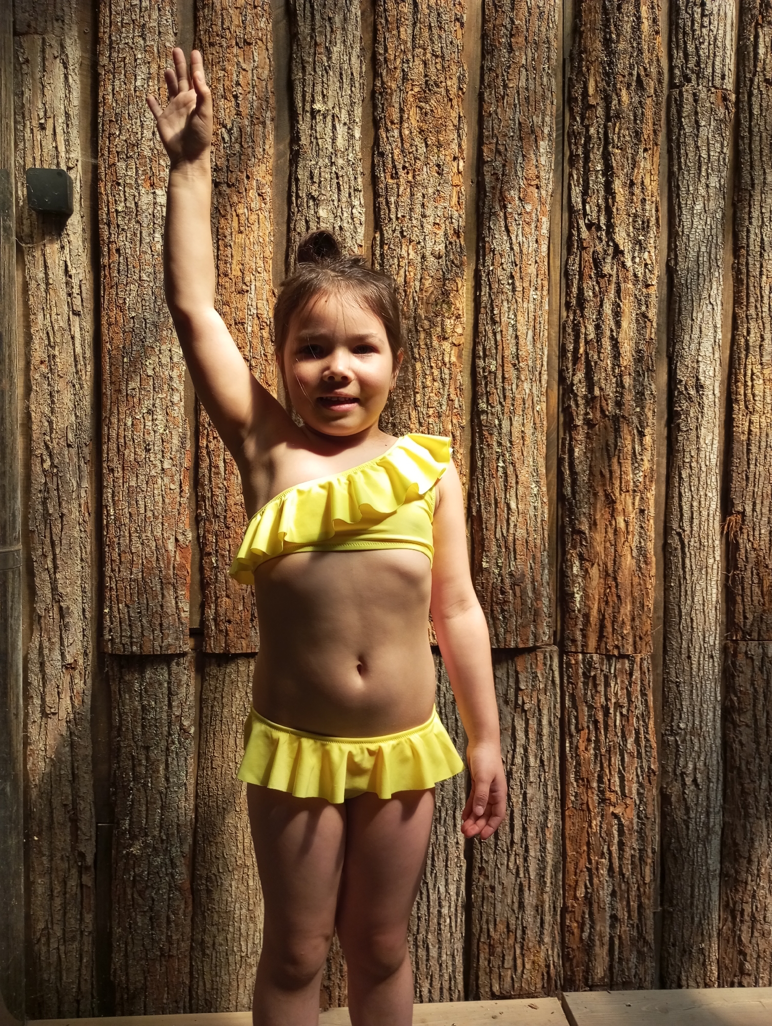 A young girl stands facing the camera in a yellow, one-shoulder swimsuit with ruffles. Her right arm is raised in the air, and she has her hair up in a bun. The background consists of vertically stacked wooden logs arranged to form a wall. The lighting indicates an outdoor setting with sunlight casting shadows. No evidence points to the location being in Izhevsk, Russia.