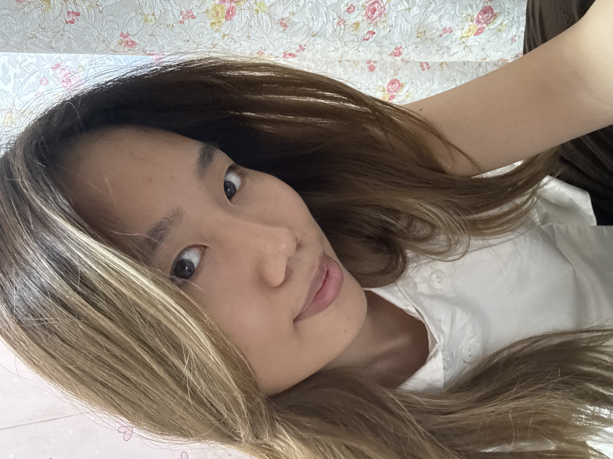 The image shows a close-up selfie of a young Asian woman with long, wavy brown hair with blonde highlights. She is wearing a white collared shirt with visible buttons. Her left eye is visible, and she has a slight smile on her lips. The background consists of two patterned wallpapers; one on the left appears to have a pink, abstract design with hearts, and the one on the right features a floral pattern with pink and yellow flowers. The lighting suggests an indoor setting, possibly a room. No information regarding her location in Shapingba District, China, is discernible from the image itself.
