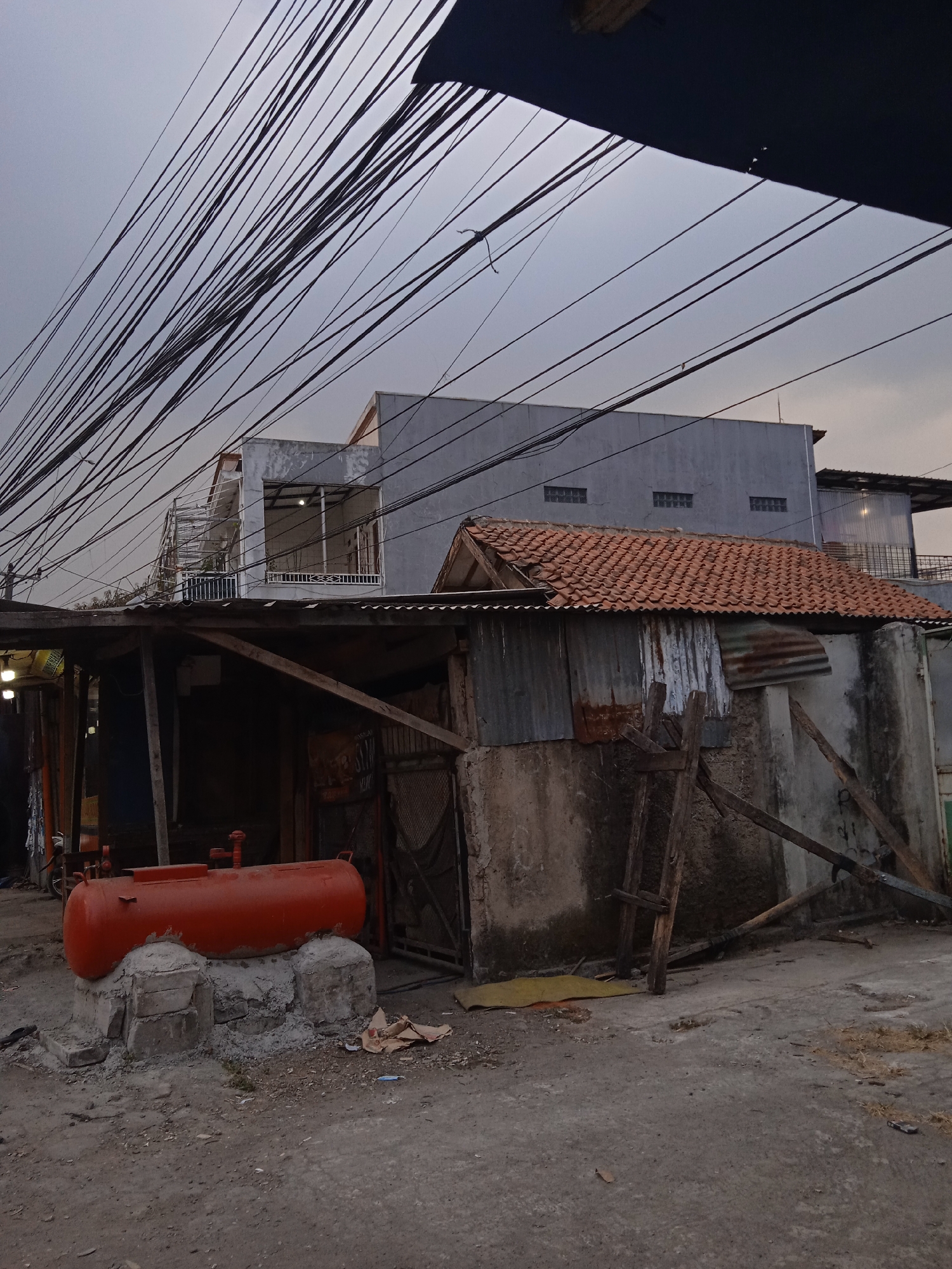 The image depicts an outdoor scene with a cloudy sky and numerous overhead power lines. In the foreground, a large, orange, cylindrical tank rests on a concrete and stone foundation. The area surrounding the tank is unpaved, with some scattered debris. Behind the tank, a low-lying structure with a corrugated metal roof and a rough concrete wall is visible. Wooden supports and framing are apparent, suggesting an unfinished or dilapidated state. Further back, a multi-story building with a grey facade and visible windows stands under the dense network of power lines. The scene appears to be in a semi-urban or industrial area in Cileunyi, Indonesia, characterized by a mixture of structures and utilitarian elements. There are no people visible in the image.