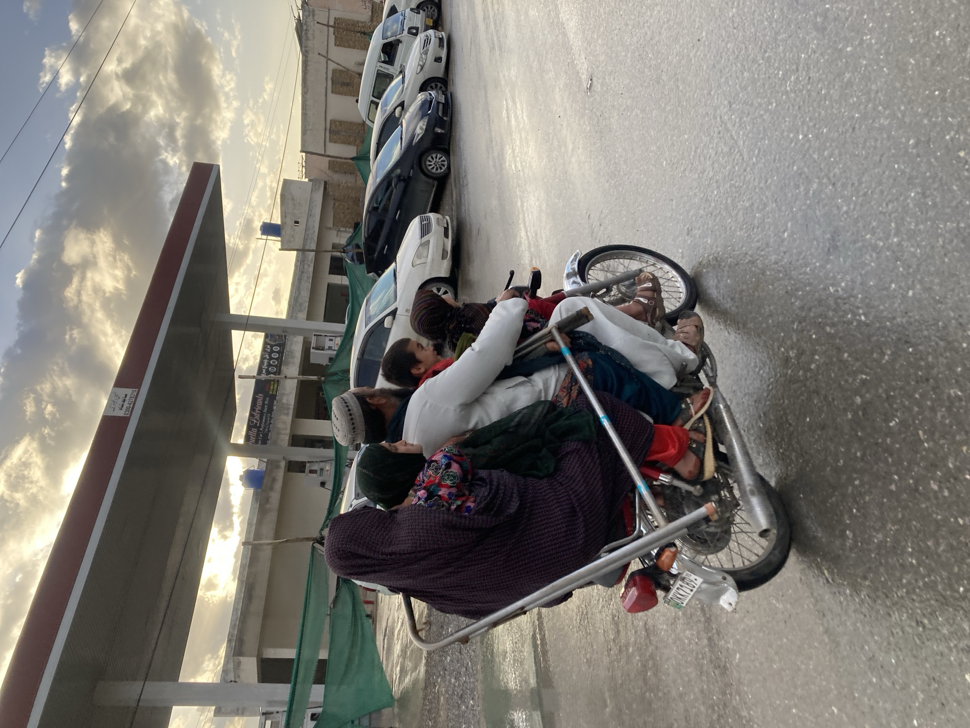 A silver motorcycle travels on a wet asphalt road in Quetta City Tehsil, Pakistan. The motorcycle transports four individuals: an adult male driver, a child seated directly in front of him, another child positioned between the driver and an adult female, and the adult female seated at the rear. The male driver wears a white shalwar kameez and a white skullcap. The child in front of him wears red clothing. The child between the driver and the female wears a green scarf and a patterned top. The adult female, positioned at the rear, is draped in a dark purple shawl over red pants. The motorcycle features a custom metal frame extending from its rear and displays the license plate "BKK 7189" on its rear fender. In the background, a fuel station structure with a prominent canopy is visible. A sign for "Quetta Lubricants" and a phone number (0300-4278510) are discernible on the station building. Several parked vehicles, including sedans and a white van, are visible on the station premises. Green mesh fencing lines the mid-ground. The sky overhead is partially cloudy, with diffused sunlight.