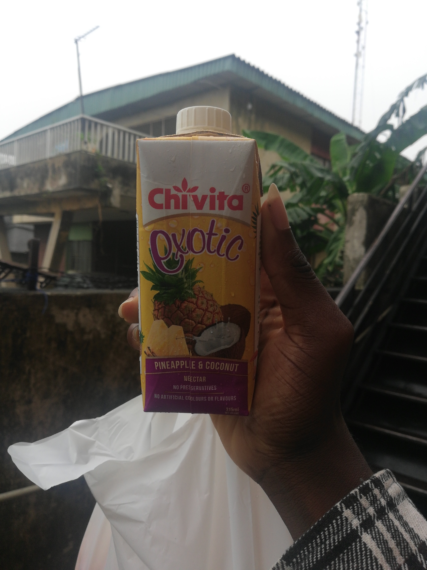 The image depicts a person holding a carton of Chivita Exotic Pineapple & Coconut juice. The background shows an urban scene, likely in Lagos, Nigeria, given the style of architecture and infrastructure. Buildings are visible, including a partially obscured one with a balcony and another structure with a rooftop antenna. A metal staircase is present on the right side. A white plastic bag is visible beneath the juice box. The overall scene appears to be a typical urban environment with overcast lighting.