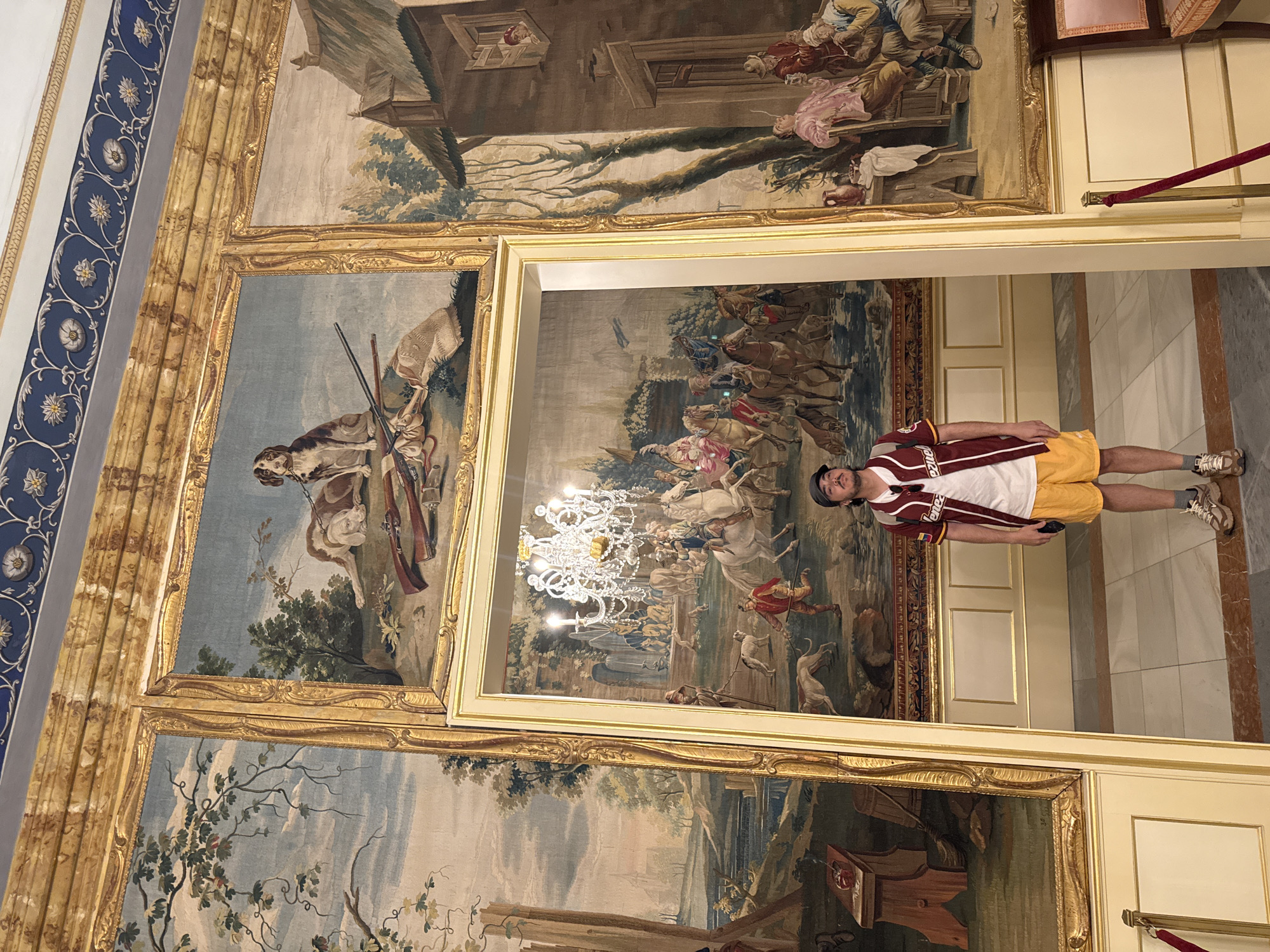A young adult male is standing in a doorway within a grand interior space adorned with large, ornate tapestries. The man, wearing a maroon and white baseball jersey over a white t-shirt, yellow shorts, and sneakers, is looking towards the camera. He has a backpack on his shoulders and is holding a phone in his right hand. The doorway he stands in reveals a glimpse of another tapestry depicting a pastoral scene with people on horseback, a dog, and a fountain, illuminated by a large chandelier. The surrounding walls are covered with expansive tapestries featuring hunting dogs, hunting rifles, and rural village scenes. The tapestries are framed by gilded borders and are mounted on walls with decorative molding and a blue and white patterned frieze along the top. The floor is tiled. This scene is located within the Royal Site of San Lorenzo de El Escorial, Spain, a complex that includes a royal palace, basilica, pantheon, monastery, and museum.