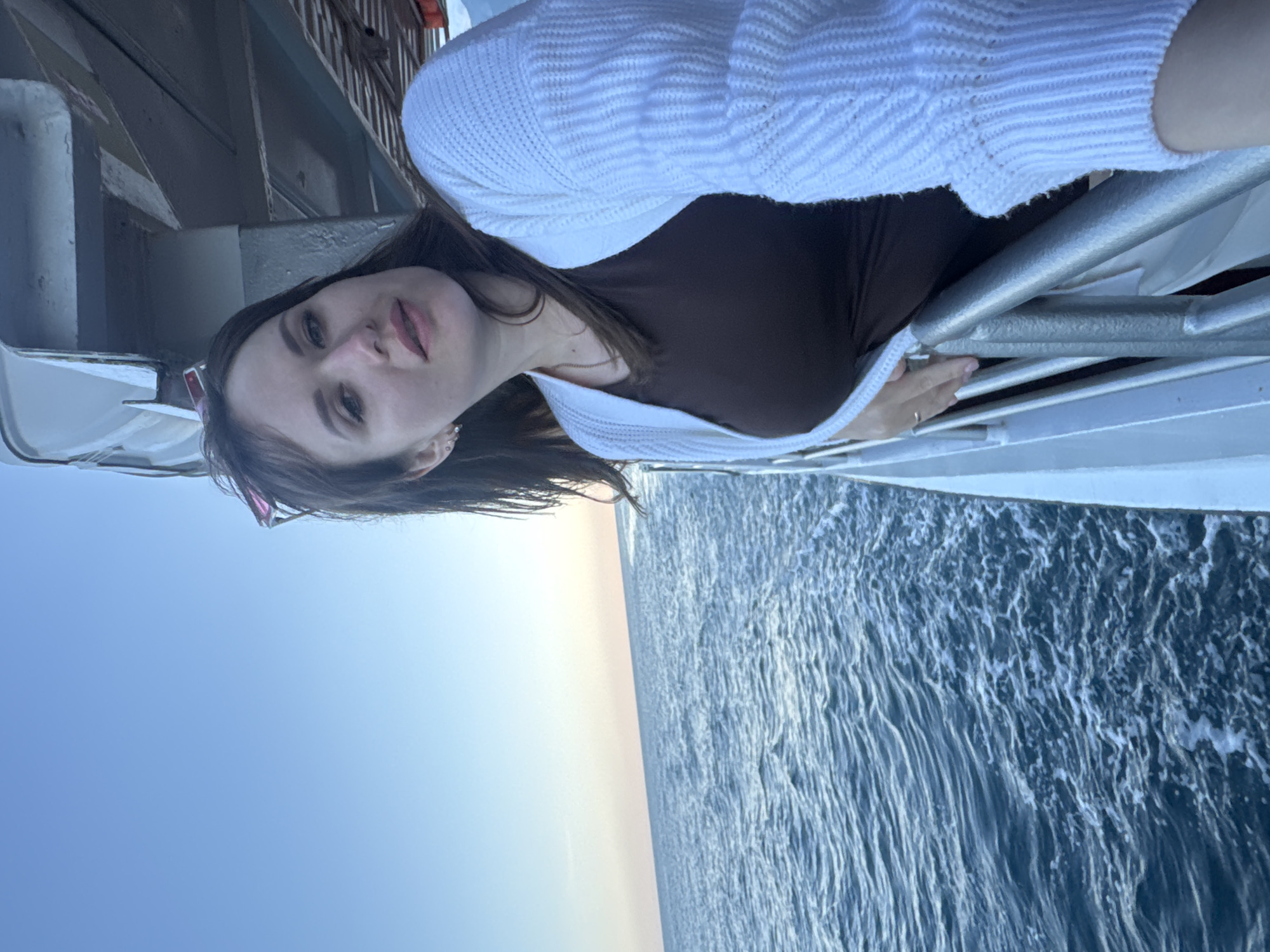 A woman is seen standing on what appears to be a boat or a ferry. She is wearing a brown top and a white cardigan, and she is holding onto a silver railing. Beyond the railing is a view of the sea. The sky is visible above the sea, showing a gradient of light blue and white. The sea itself is a dark blue with white waves.
The location appears to be Гаспринский поселковый совет, Ukraine, given the presence of water and coastal features. The woman is the primary subject, and the sea and sky form the backdrop.