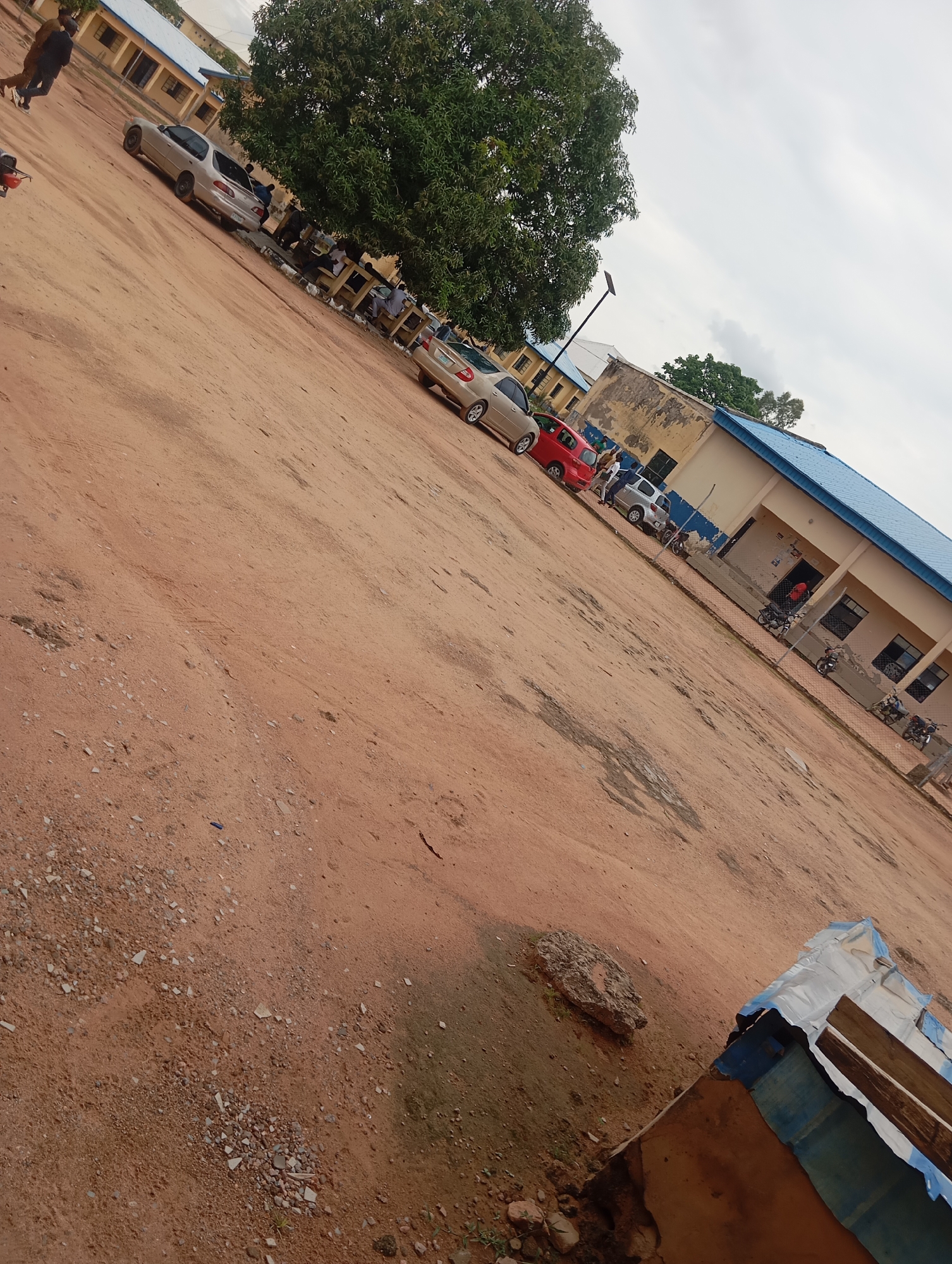 This image depicts an outdoor scene, likely within the city of Jalingo, Nigeria. In the background, a light-colored building with a blue roof is visible. Several cars are parked in the dirt lot in front of the building, including a silver sedan, a red hatchback, and another silver sedan. A large tree with green foliage stands to the left of the building, casting shadows on the ground. A few individuals are walking in the foreground and near the cars. The ground is covered with reddish-brown dirt and scattered gravel. A makeshift shelter constructed with blue tarp and wooden planks is present in the lower right corner of the image. The sky is overcast with clouds.