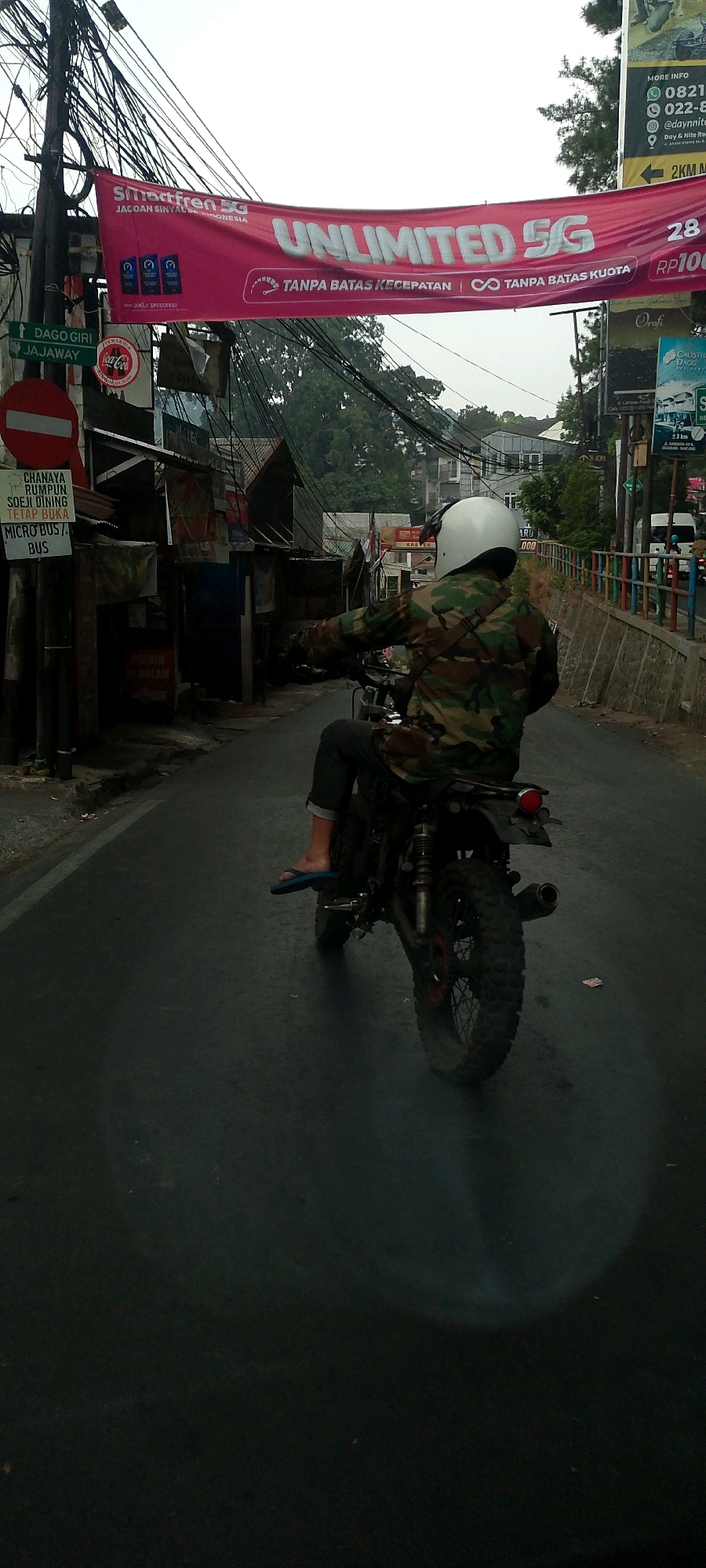 A person wearing a camouflage jacket and a white helmet rides a motorcycle on a street in Bandung City, Indonesia. The person is in the foreground, viewed from behind, with their left leg resting on the motorcycle's footrest and wearing blue flip-flops. The motorcycle is dark-colored, with a visible exhaust pipe and knobby tires. In the background, a large pink banner hangs across the street, advertising "Smartfren 5G" and "UNLIMITED 5G TANPA BATAS KECEPATAN | TANPA BATAS KUOTA." There are also various signs and posters on poles and buildings, including directional signs pointing to "DAGO GIRI" and "JAJAWAY," as well as advertisements for Coca-Cola. A traffic sign prohibiting entry is also visible. The street appears to be a two-way road with a slight incline, lined with trees and buildings on either side. The sky is overcast, suggesting it might be a cloudy day. The overall scene depicts a street view in a city, with elements of commerce and transportation.