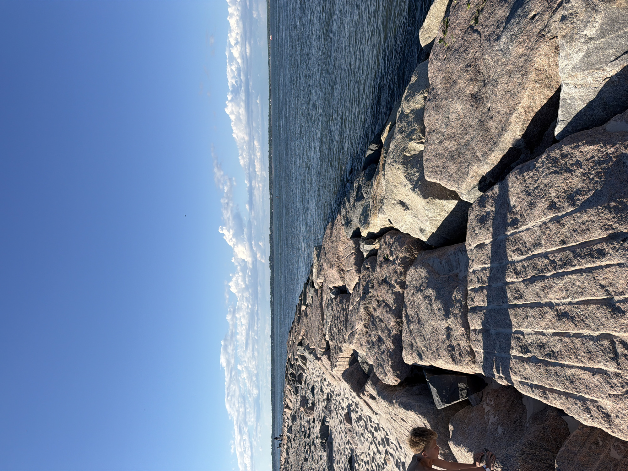 The image depicts a sunny day at a beach in Hampton Bays, New York. A person, a young boy, is seen on the left side of the frame, bending down on a sandy area next to a large stone jetty. The jetty, composed of massive pink and grey granite rocks, extends into the dark blue ocean. The water is calm with small ripples. In the distance, a thin strip of land with trees and some structures is visible, bordering the horizon. The sky above is a clear, bright blue with scattered white cumulus clouds. The sandy beach is visible to the left of the jetty, with footprints imprinted on its surface.
