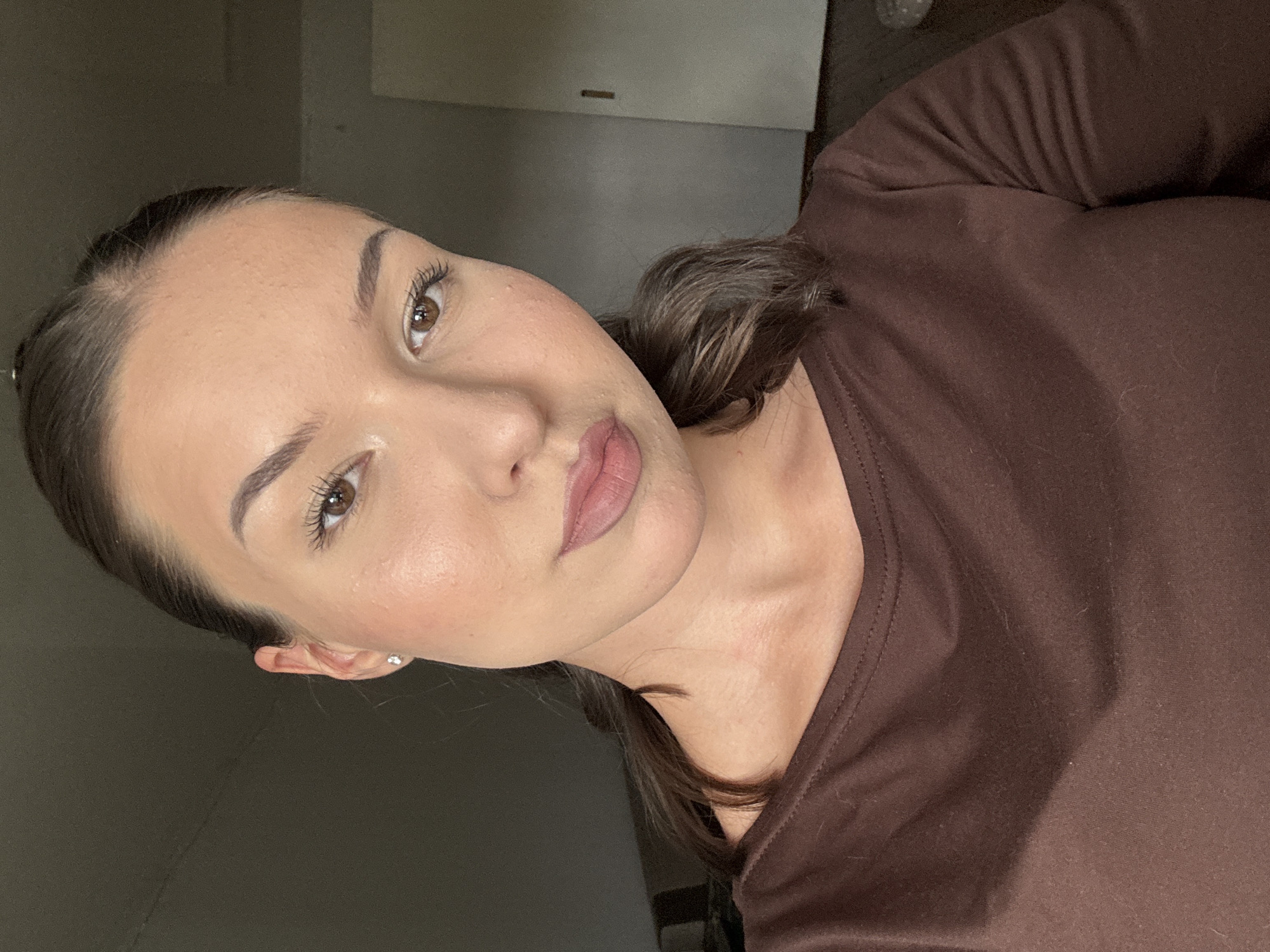 A woman is looking at the camera, positioned slightly off-center to the right. She has dark hair pulled back, visible makeup, and is wearing a brown long-sleeved shirt. The background is a dimly lit room with a dark wall on the left and a lighter wall with a door on the right. The floor is visible in the lower right corner. The scene is likely indoors, possibly in a residential setting, within the city of Inskoy, Russia.