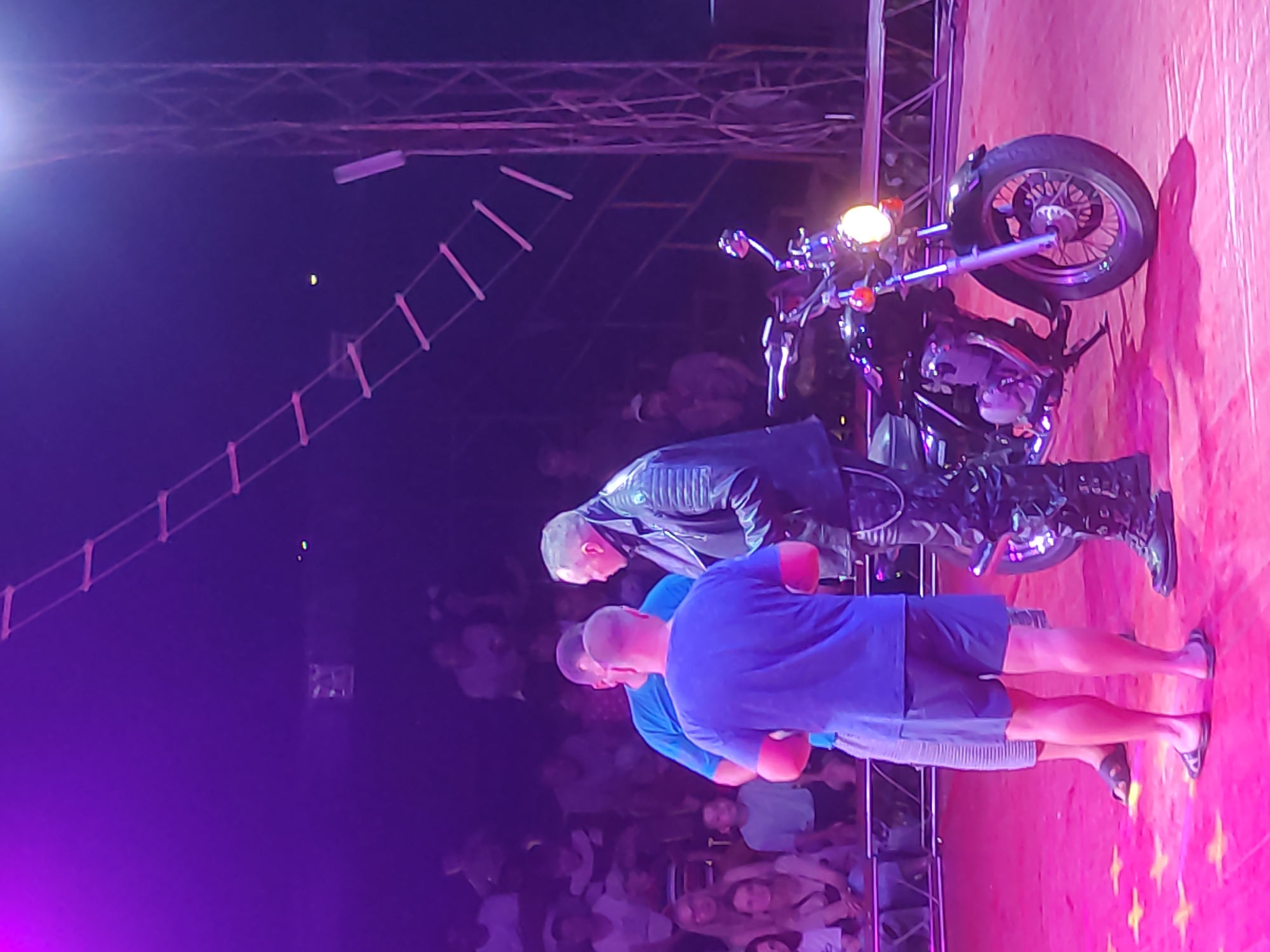 In the city of Mahilyow, Belarus, a motorcycle stunt is taking place in a circus tent. Three men are visible on the pink-carpeted stage. Two men in casual clothing are facing a third man in a black leather jacket and pants, who is standing next to a black motorcycle. The audience is seated in the background, illuminated by purple lights. A ladder-like structure hangs from the ceiling, and a metal tower stands to the right. The motorcycle has a lit headlight, and the stage floor is decorated with yellow stars.