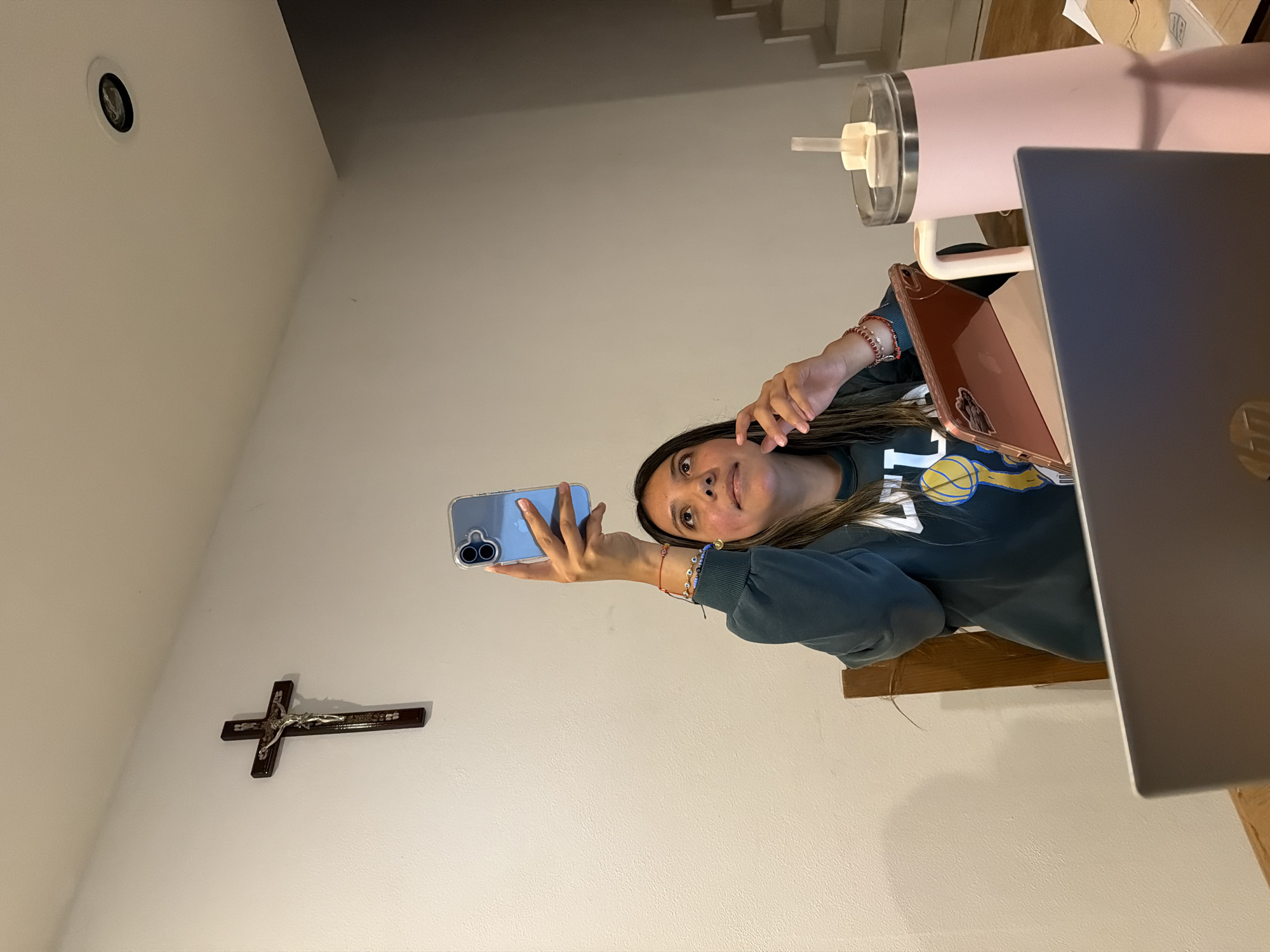 A fair-skinned woman with dark hair and brown eyes holds a blue smartphone up in her right hand, apparently taking a selfie, in what looks like a residence in San Andrés Cholula, Mexico. She is wearing a blue-green sweatshirt with yellow and white graphics. On the wall to her left hangs a crucifix. A light fixture is also visible in the upper-left corner. To the right of the woman is a laptop and a pink cup on a wooden surface, presumable a desk or table. Also visible is a brown tablet with a clear case. The woman's left wrist is adorned with multiple bracelets.