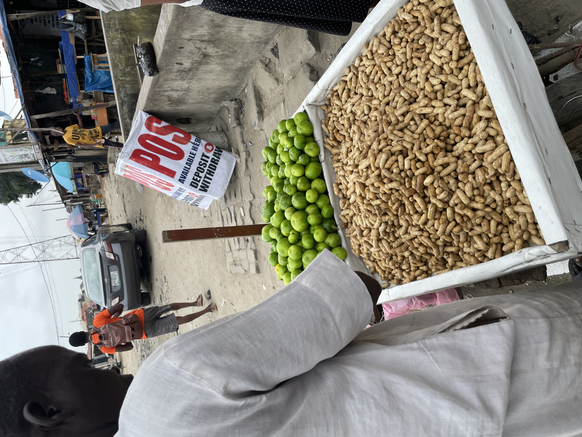 In Ado, Nigeria, a street vendor displays a plentiful harvest of peanuts in a large white container, situated next to a brimming bin of vibrant green limes. The vendor, partially obscured by a light-colored tunic, stands between the produce and a paved area littered with debris. In the background, a dark sedan with a license plate reading "APP 811FL" is parked on the dusty street. A man in an orange shirt and backpack walks away from the camera. Several stalls with blue tarps and signage are visible further down the street, suggesting a bustling market environment. A large white banner with red lettering that reads "POS AVAILABLE HERE DEPOSIT & WITHDRAW" leans against a concrete wall, indicating financial services are offered. The sky above is overcast.
