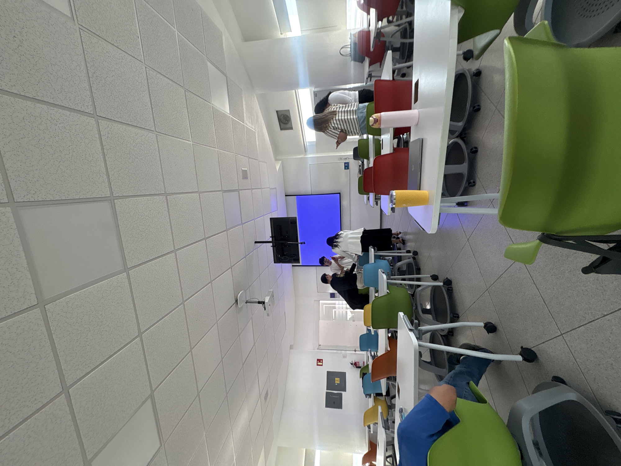 A classroom in Puebla, Mexico, is shown, with tables and chairs arranged in rows. A projector screen displays a blue image at the front of the room. Several people are present in the room, including students and possibly an instructor. Some individuals are sitting at the tables, while others are standing near the screen. The ceiling is a drop ceiling. The walls are white. There are emergency signs on one wall.