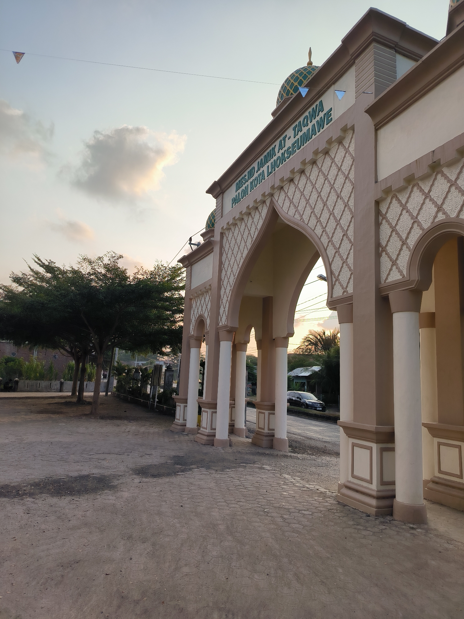 The image shows the entrance to the At-Taqwa Mosque in Paloh, Lhokseumawe, Indonesia. The mosque's entrance features arches with a decorative pattern and beige-colored columns. A green dome is visible on top of the building. The sky is overcast with some clouds, suggesting it may be early morning or late afternoon. Trees are visible to the left of the entrance, and a car is parked on the street in the background. The ground in front of the mosque is paved with cobblestones. The scene is devoid of people.