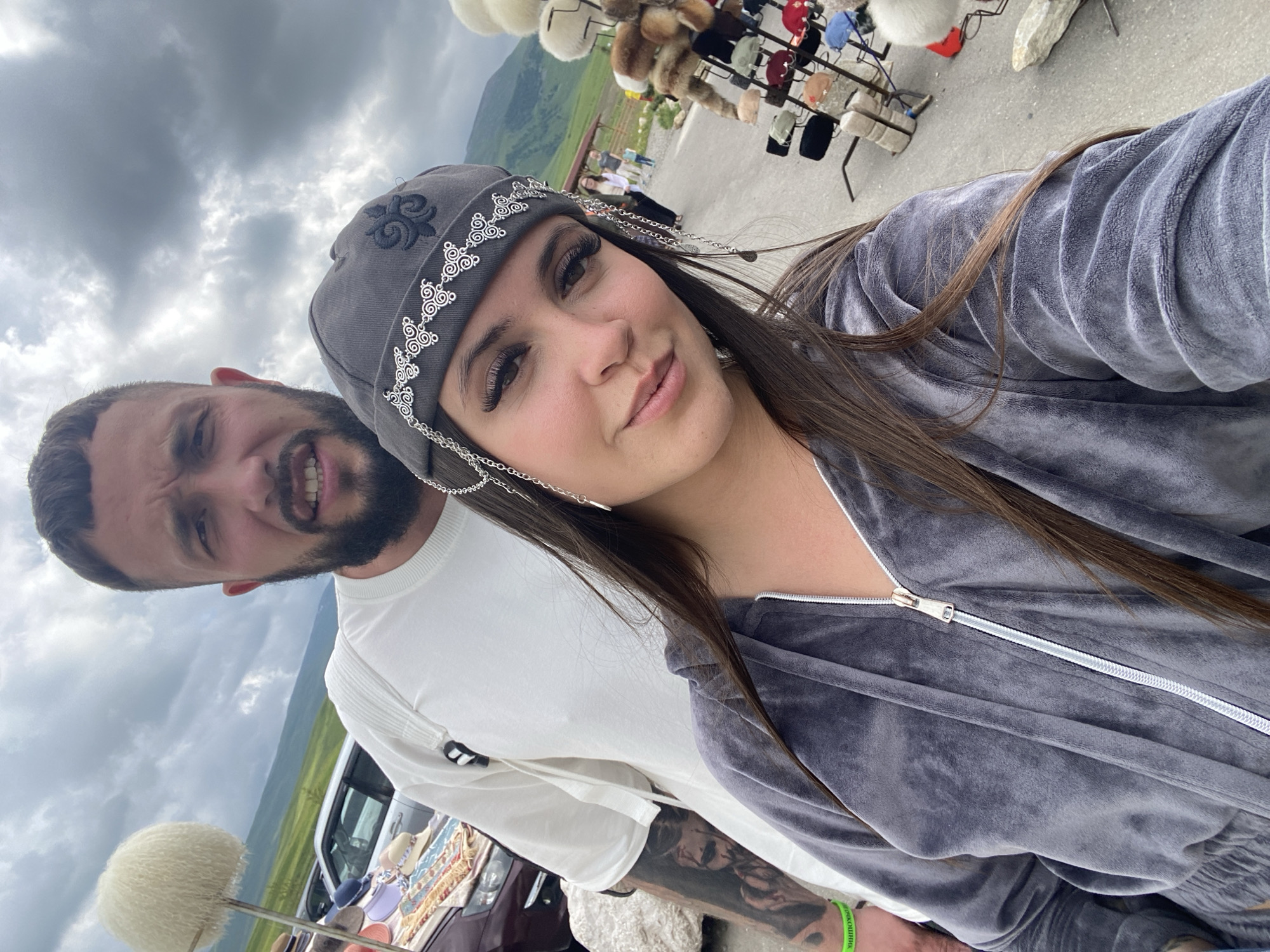 A man and a woman are posing for a selfie in an outdoor market setting. The man, positioned behind the woman, is wearing a white t-shirt and has a beard. The woman, in the foreground, is wearing a grey velvet tracksuit and a grey hat adorned with silver chain embellishments and embroidery. She has long brown hair and prominent eyelashes. The background shows a display of various hats, cars, and a hilly, overcast landscape, indicative of a rural or mountainous region. The location is identified as Tsalkita, Russia, implying it's a market or tourist spot within that area.