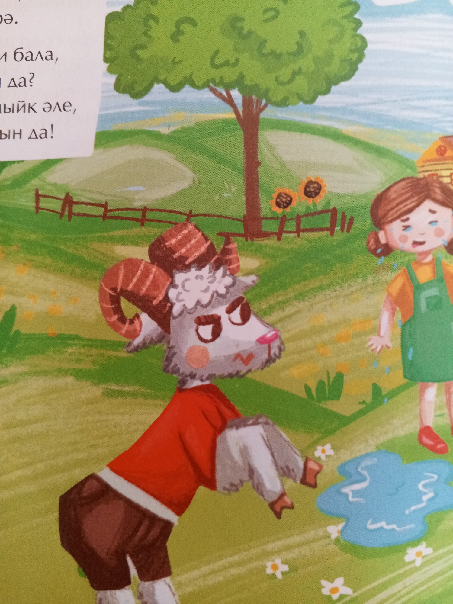 The image depicts a whimsical, cartoon-style illustration with a bright, grassy landscape under a pale blue sky. In the foreground, a goat with large, curved horns, wearing a red shirt and brown pants, stands facing left with a grumpy expression. To the right, a young girl with brown pigtails and a green apron over a yellow shirt is crying, with tears streaming down her face. She stands near a blue puddle on the grass. In the background, a large, leafy green tree stands with two sunflowers planted near its base, partially enclosed by a simple wooden fence. A small yellow building with a red roof is visible further in the background. The overall scene suggests a moment of childhood distress or conflict, rendered in a playful, illustrative style. The mention of Chistopol, Russia, in relation to this image does not correspond to any visual elements within the illustration itself.