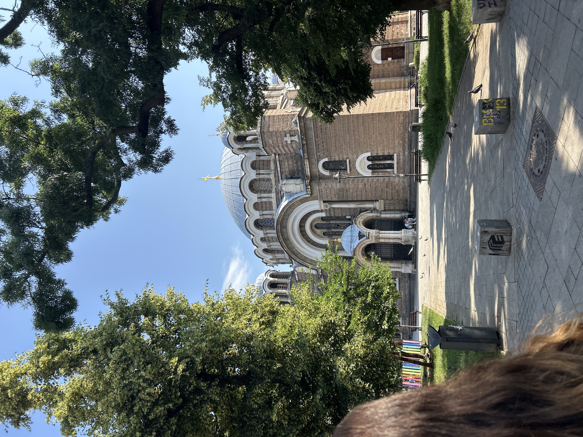 The image shows the Alexander Nevsky Cathedral in Sofia, Bulgaria, on a sunny day. The large, ornate cathedral, built of brick, features multiple domes, the largest of which is covered in copper and topped with a golden cross. The facade has arched windows and doorways. The cathedral is framed by lush green trees, with some branches extending into the foreground.
In the foreground, a person's brown hair is visible on the left, obscuring part of the scene. A paved walkway leads towards the cathedral, with a trash can and several concrete blocks, some with graffiti, placed along the path. Pigeons are scattered across the walkway and grass. Benches are visible in front of the cathedral, and a few people are seated on them. In the background, behind the cathedral, a clear blue sky is visible with a few wispy clouds. Colorful decorative posts are also visible to the left of the walkway.