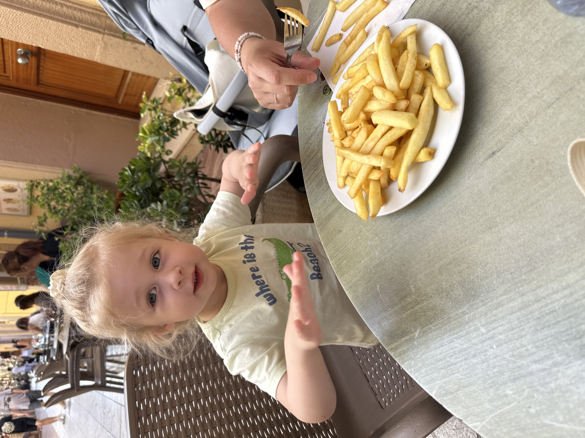 In Sant Feliu De Guíxols, Spain, a young blonde child with blue eyes sits at a table outdoors, facing the camera with their hands outstretched towards a plate of french fries. The child wears a pale yellow t-shirt with the text "where is the Beach?" and an illustration of a crocodile. A person's hand, wearing a ring and bracelet, is visible on the right, holding a fork with a french fry, about to offer it to the child. The table is round and has a light green, textured surface. In the background, there are other tables and chairs, a stroller, potted plants, and a wooden door. The overall setting appears to be a restaurant patio on a street with other people in the distance.