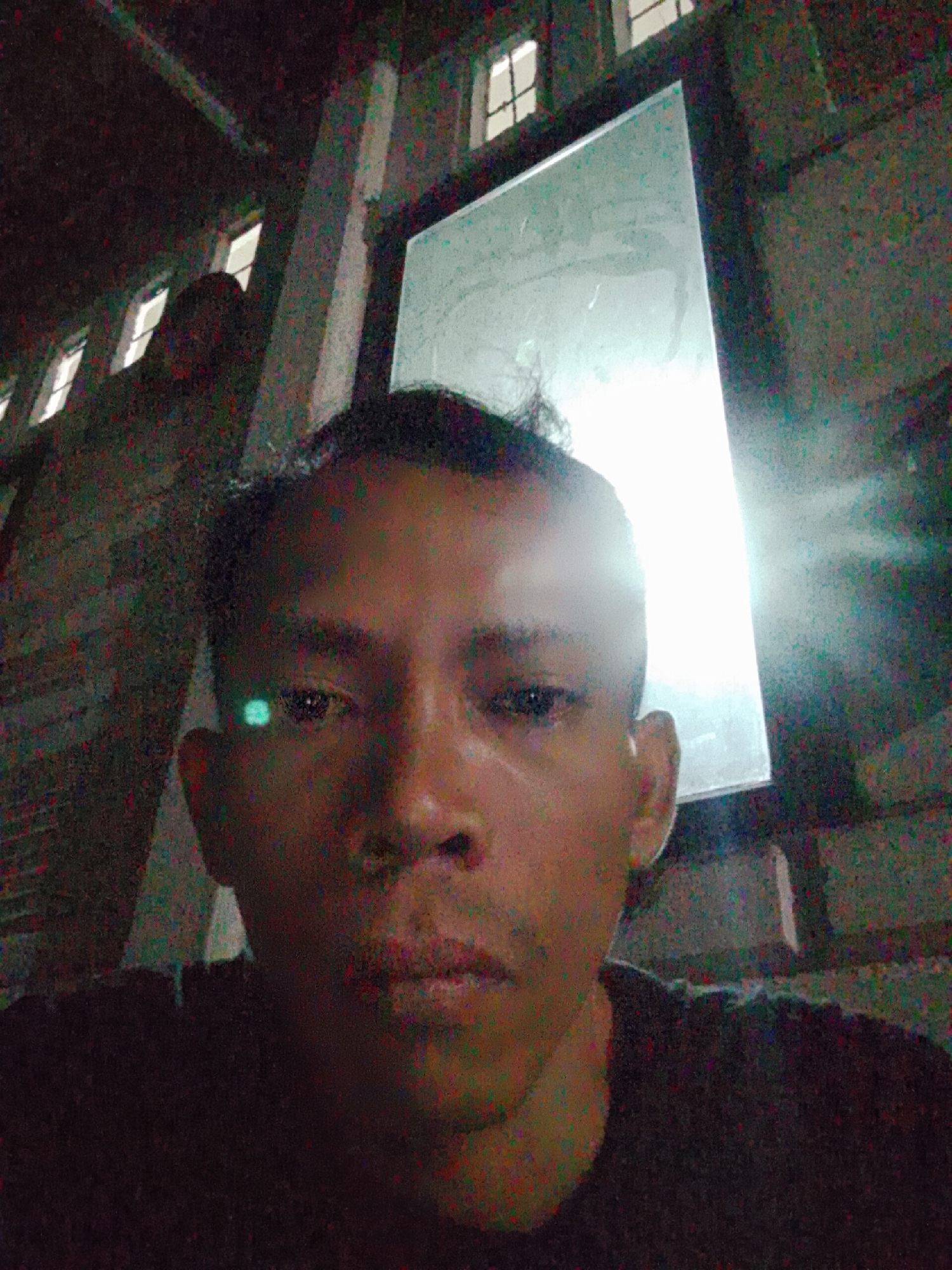 A man is standing outdoors, likely in Bukit Ketok, Indonesia, based on the location provided. He is wearing a dark shirt. Behind him, there is a structure with windows, one of which is brightly illuminated. The image quality is low, with noticeable graininess.