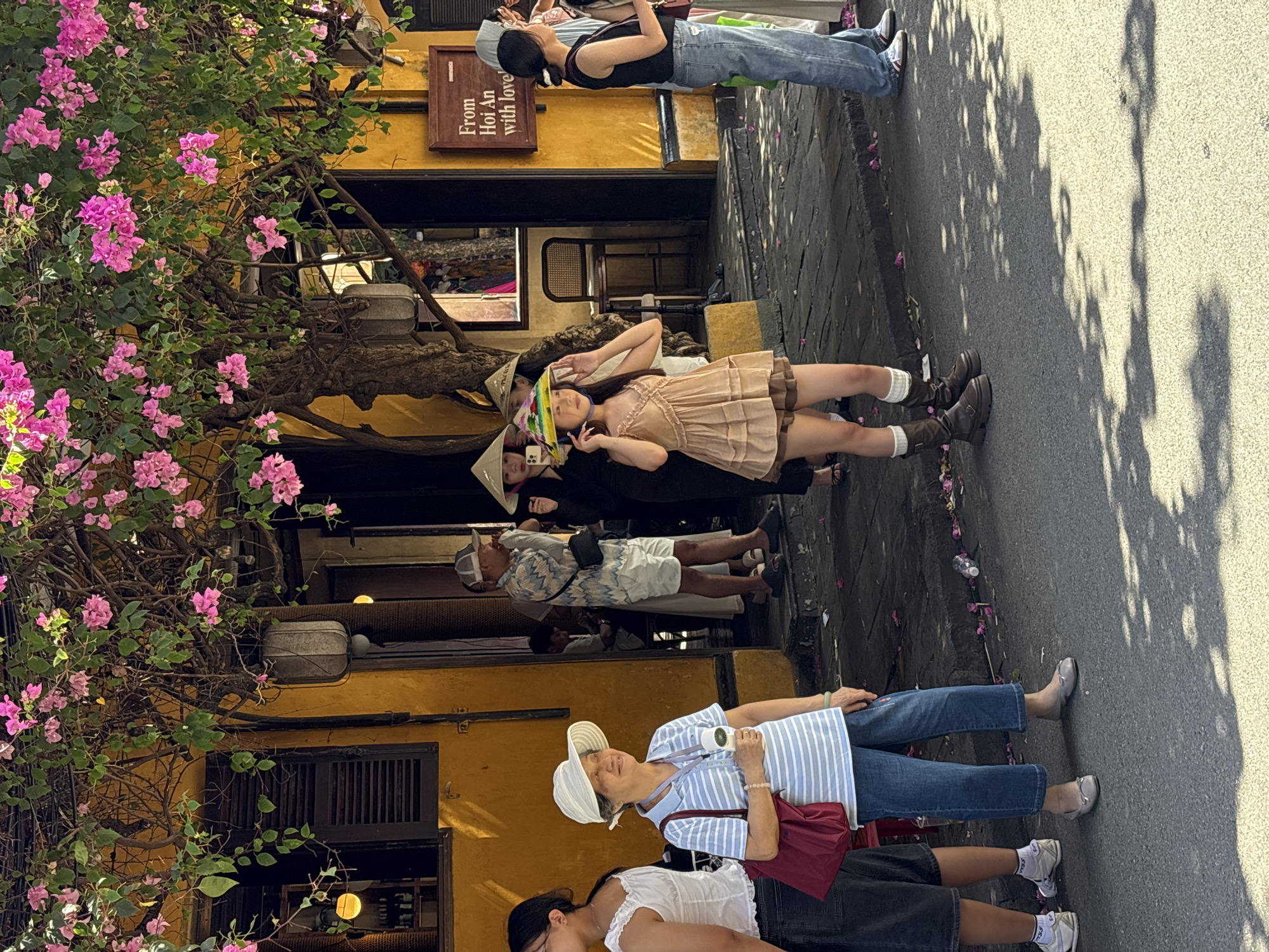 This image depicts a street scene in Hội An, Vietnam, with a focus on people and the surrounding architecture. The building in the background is painted a vibrant yellow and is adorned with an abundance of pink bougainvillea flowers cascading from above. A sign on the building reads "From Hoi An with love." Several individuals are visible on the sidewalk and street.
In the foreground, on the left, a woman wearing a white hat and a blue and white striped polo shirt is holding a small handheld fan. She has a red bag slung over her shoulder and is wearing blue jeans and grey ballet flats. To her left, partially visible, is another person in a white top and dark shorts, wearing white sneakers and socks.
Further back, on the sidewalk, a young woman in a light brown, ruffled dress and white socks with brown boots is holding her hand to her conical hat, adjusting it. Behind her, other individuals wearing traditional conical hats are also visible.
To the right, a person in a black tank top and blue jeans is standing on the sidewalk, looking away. Another person in a light-colored outfit is partially visible to their right. The street itself is paved and shadowed by the trees and buildings. Fallen pink petals are scattered on the sidewalk and street. A plastic water bottle is visible on the edge of the sidewalk. The overall atmosphere suggests a sunny day in a lively urban setting.