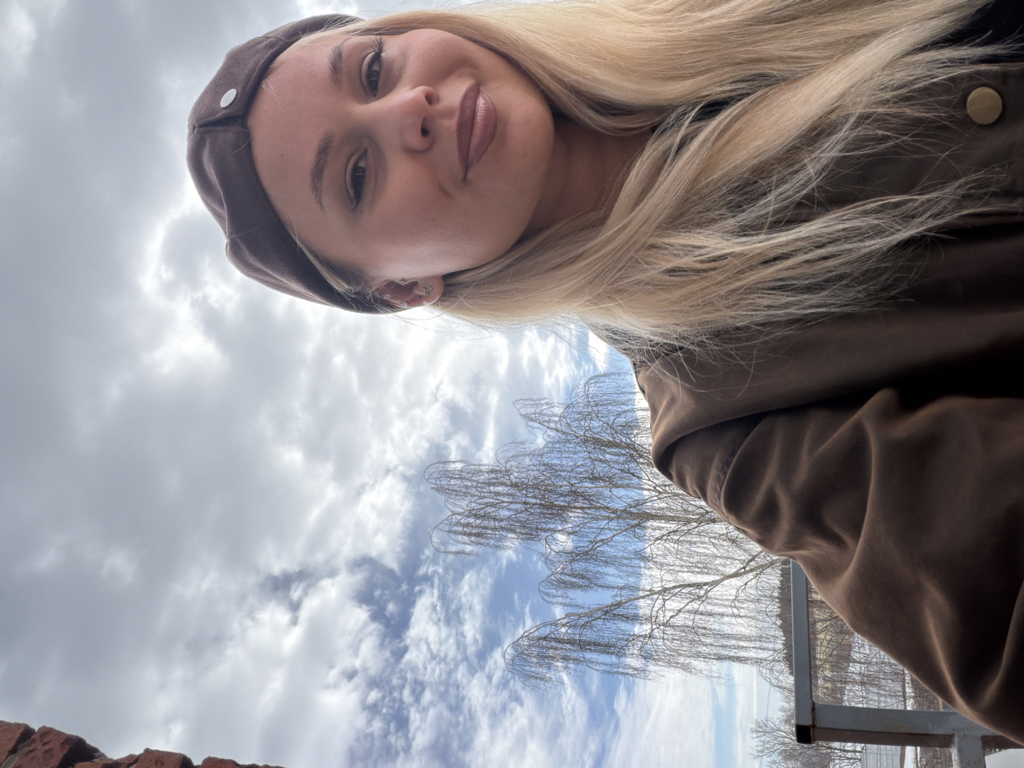 A female individual is depicted in a selfie, taken outdoors in Sarapul, Russia. The subject is a young adult with long blonde hair, fair skin, and multiple silver piercings on her left ear. She wears a brown beret-style hat featuring a single silver button and a brown outer garment. Her lips are accentuated with a light brown lip color, and she presents a subtle smile while gazing directly at the camera. The background consists of a partially cloudy sky with visible blue patches. To the immediate left, a section of a reddish-brown brick wall is visible. Behind the subject, a large deciduous tree with bare, drooping branches stands prominently, indicating late autumn, winter, or early spring. Below the tree, a grey metal railing is present, overlooking a distant landscape with sparse vegetation and light-colored ground, consistent with a semi-urban or suburban setting within Sarapul.