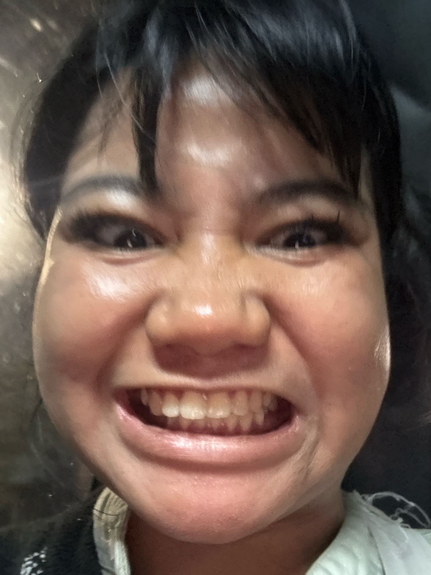 A close-up, full-face, selfie-style portrait of a young East Asian woman with dark hair, wrinkled eyes, and an exaggerated, wide grin revealing her teeth. The woman's cheeks are puffed out, and her eyes are squinted. She appears to be wearing makeup, including prominent eyelashes. The background is dark and out of focus, with hints of reflective surfaces, suggesting an indoor setting, possibly at night. The image is grainy and has a somewhat distorted fisheye lens effect. The image is located in North Jakarta, Indonesia.
