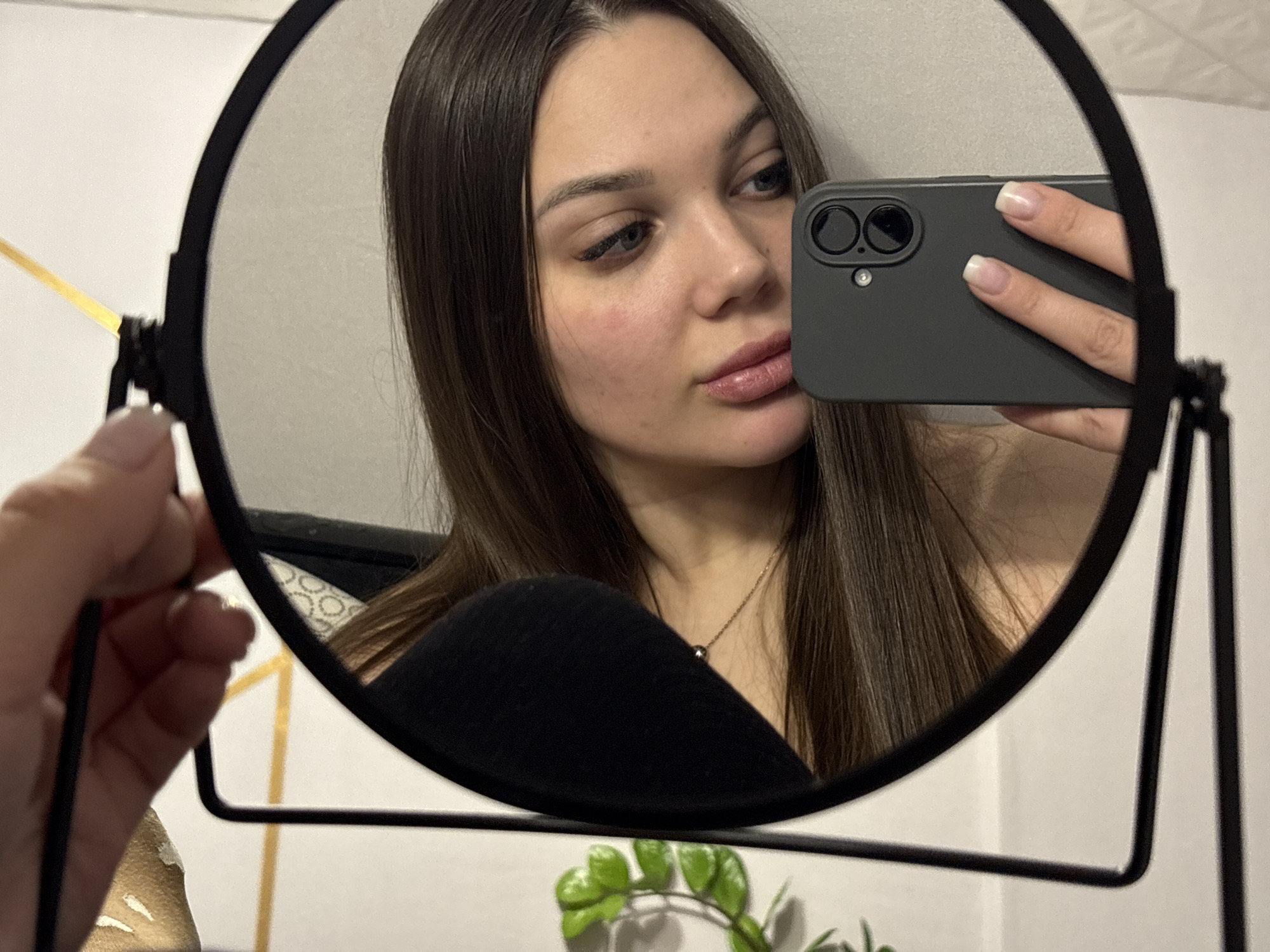 A young woman takes a selfie in a round, black-framed mirror. She holds a smartphone with a gray case in her right hand, partially obscuring her face. Her hair is long, straight, and dark brown. She has fair skin and is wearing subtle makeup. She is wearing a black top and a necklace. The background includes a white wall with a gold geometric design, a small green plant and a person holding the mirror. It is not possible to confirm the location in the city of Chaikovsky, Russia without additional information.