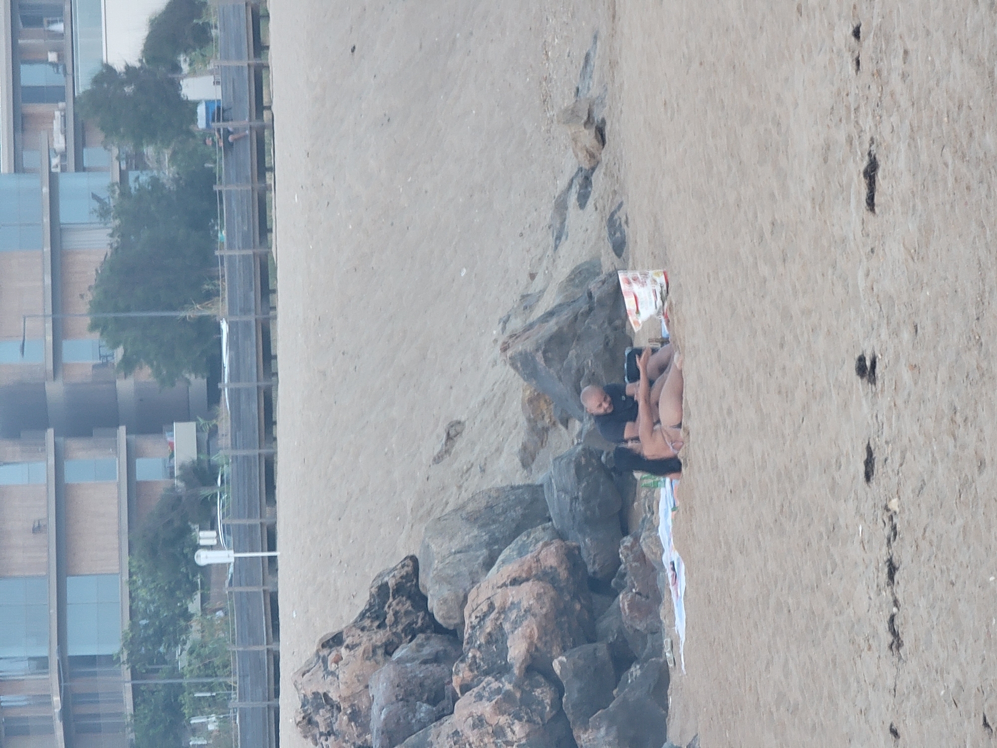 The image depicts a beach scene in Quarteira, Portugal, with a focus on a couple sitting on the sand. They are positioned near a cluster of large rocks, with a sandy slope rising behind them. A modern building complex with large glass windows is visible in the background, along with some trees and a wooden boardwalk. The couple appears to be relaxing, with one person lying down and the other sitting up. There are some items on the sand near them, including a blanket and a bag. The lighting suggests it is daytime, possibly overcast or late afternoon.
