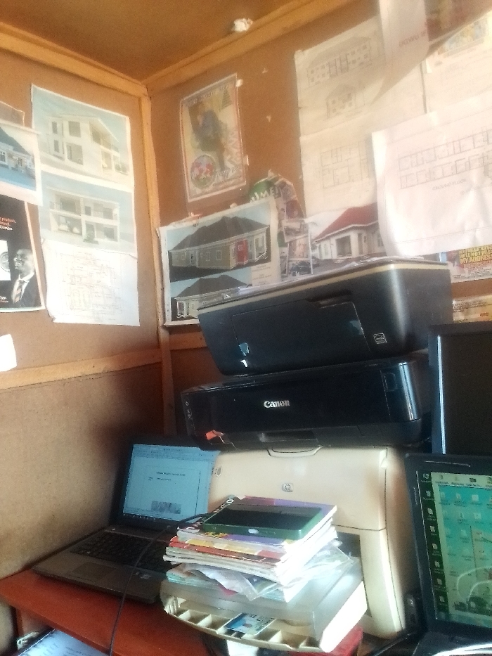 The image shows a room in Auchi, Nigeria (latitude: 7.062, longitude: 6.273). The room is a small office, with corkboard walls covered with architectural drawings and diagrams. In the corner, there are three printers stacked on top of each other. The top printer is black, the middle printer is black and says "Canon" on it, and the bottom printer is white. There are two laptops on the desk, a black one on the left and a black one on the right. The black laptop on the left is open and a document can be seen on the screen. The black laptop on the right is also open and a map can be seen on the screen. There are several books on the desk, stacked on top of the bottom printer. A power cord for the laptop is also on the desk. The room is well-lit. There is no one visible in the image.