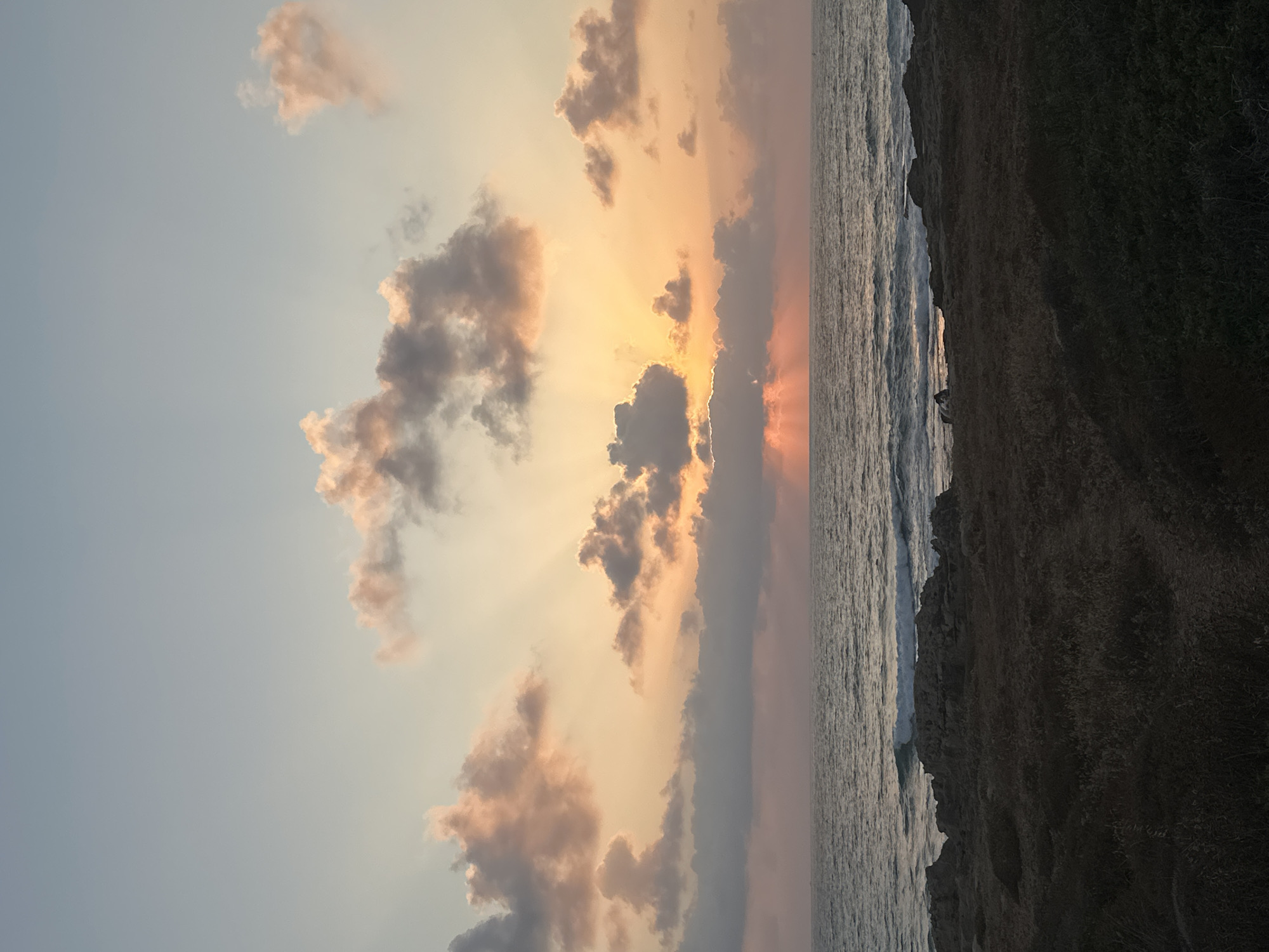 The image depicts a serene sunset over the Mediterranean Sea on the coast of Mate Asher Regional Council, Israel. The sky is a canvas of soft hues, with a pale blue transitioning into a warm orange and pink as the sun dips below the horizon. Rays of light emanate from behind scattered clouds, some tinged with pink and others a soft gray. The sea is calm, with gentle waves lapping against a rocky shoreline covered in low-lying vegetation. The foreground consists of dark, textured ground, suggesting a natural, uncultivated area. The overall atmosphere is peaceful and tranquil.