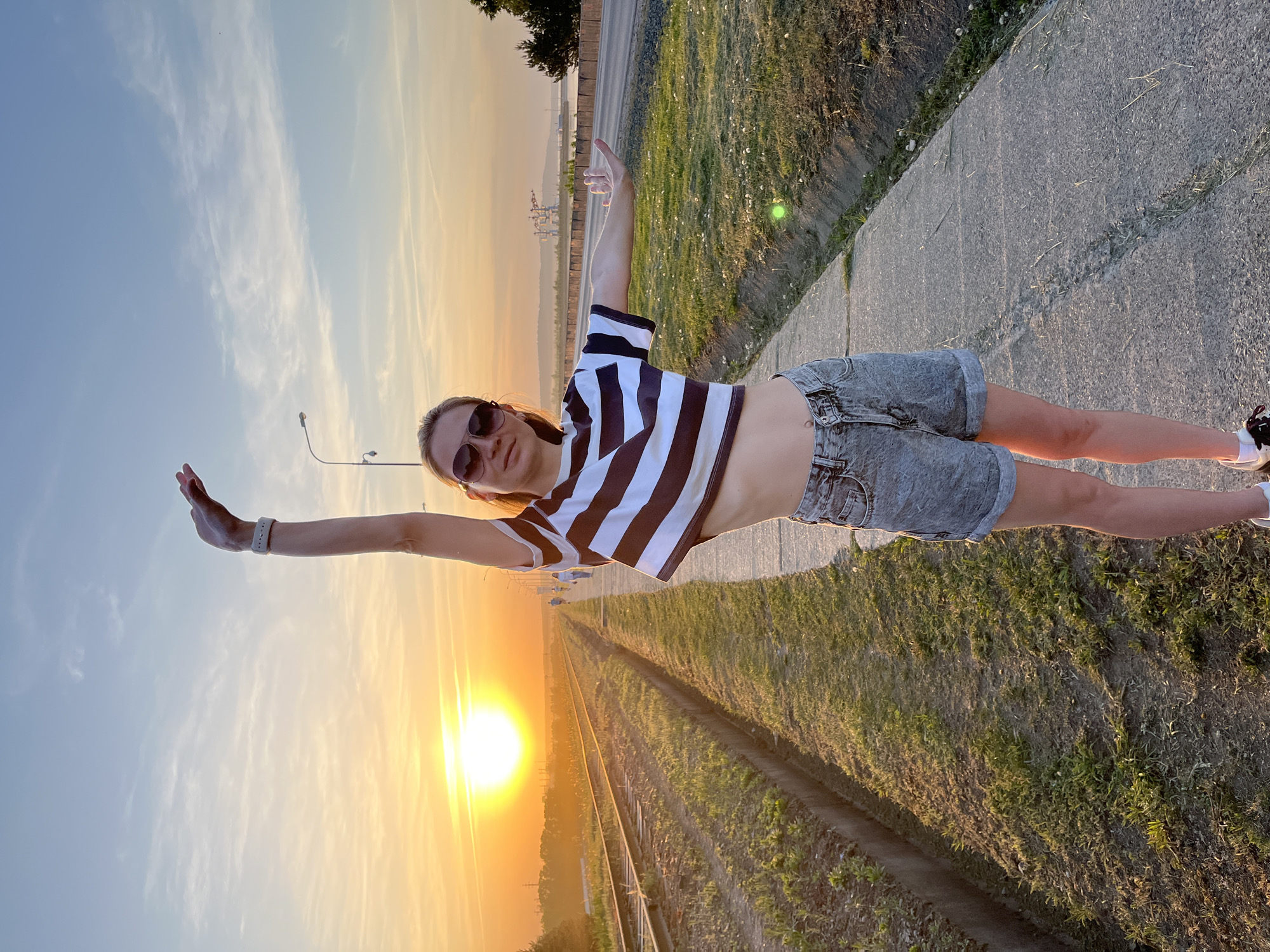 A young woman in sunglasses and a black and white striped shirt is posing on a concrete path next to railroad tracks at sunset in Chaikovsky, Russia. She is wearing denim shorts and white sneakers. One arm is extended towards the setting sun, while the other arm is extended towards the grassy area to her side. A road and a line of trees are visible in the background.
